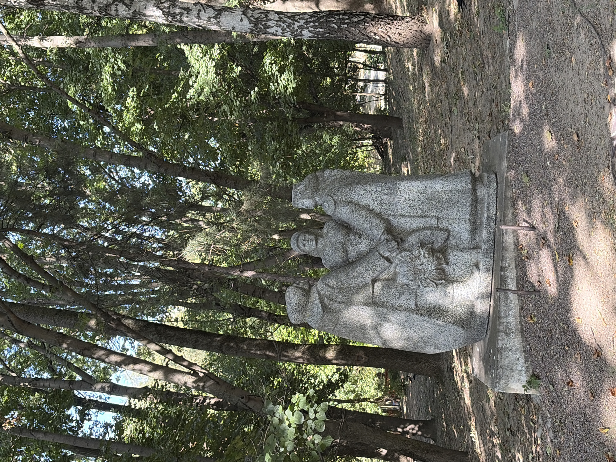 The image shows a stone sculpture of three figures in what appears to be a park setting. The sculpture is a single piece of stone, depicting three individuals standing close together, possibly holding hands or a bouquet of flowers at the center. They are dressed in clothing that suggests a historical or traditional style, with cloaks or robes. The figures are rendered in a somewhat abstract, blocky style. The sculpture is situated on a concrete base, which is itself surrounded by gravel and fallen leaves.
The background consists of a dense forest with tall trees, their branches creating a canopy that casts dappled shadows on the ground. Sunlight filters through the leaves, illuminating parts of the scene. The ground is covered in a mixture of soil, gravel, and dry leaves, typical of an outdoor park or woodland area. There are no visible signs of the city of Chișinău, Moldova, in the image itself, nor is there any explicit information that would pinpoint its exact location within the city. The overall impression is one of a quiet, natural environment with a prominent, solitary sculpture.