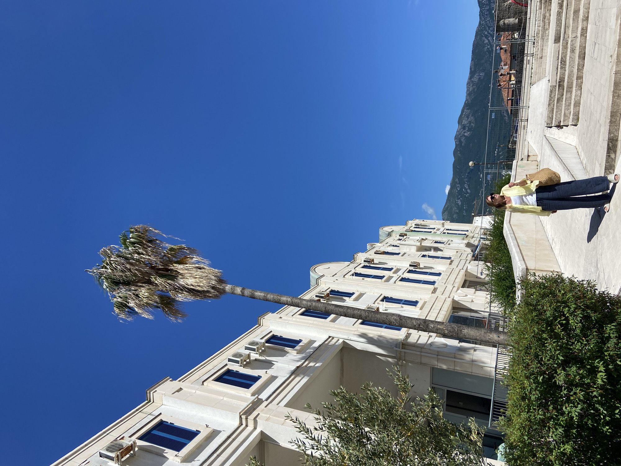 The image shows a low-angle view of a white building with blue-shuttered windows and attached air conditioning units. A palm tree stands to the left. A woman in a yellow jacket and dark pants stands on a white stone staircase to the right, holding a straw bag. Mountains are visible in the background under a clear blue sky. The scene is located in Budva, Montenegro, identified by the architectural style of the building and surrounding terrain.