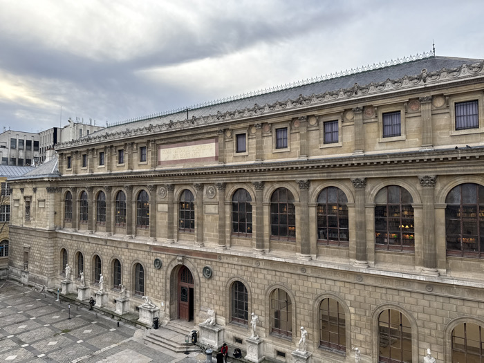 The image depicts an elevated view of a section of the Bibliothèque Nationale de France, Richelieu site, located in Paris, France. The central subject is a multi-story stone building, constructed from light-colored cut stone, featuring classical architectural elements.
The facade presents three primary levels. The ground floor and first floor above it both feature large, arched windows set between decorative pilasters. Interior lighting is visible through some of these windows, illuminating what appear to be bookshelves. A prominent stone cartouche above the first-floor windows displays the inscription "BIBLIOTHÈQUE NATIONALE DE FRANCE". The uppermost visible floor has smaller, rectangular windows. The roofline is ornate, with a cornice, balustrade, and a dark, possibly slate, pitched roof adorned with decorative finials.
At the base of the building, in the foreground, is a paved courtyard with a geometric tile pattern. A central entrance with stone steps leads into the building. Two individuals, appearing small in scale, are standing near the base of these steps. A series of white classical statues on pedestals are spaced along the building's base within the courtyard. In the far background, to the left, less ornate, modern buildings are partially visible under an overcast sky.
