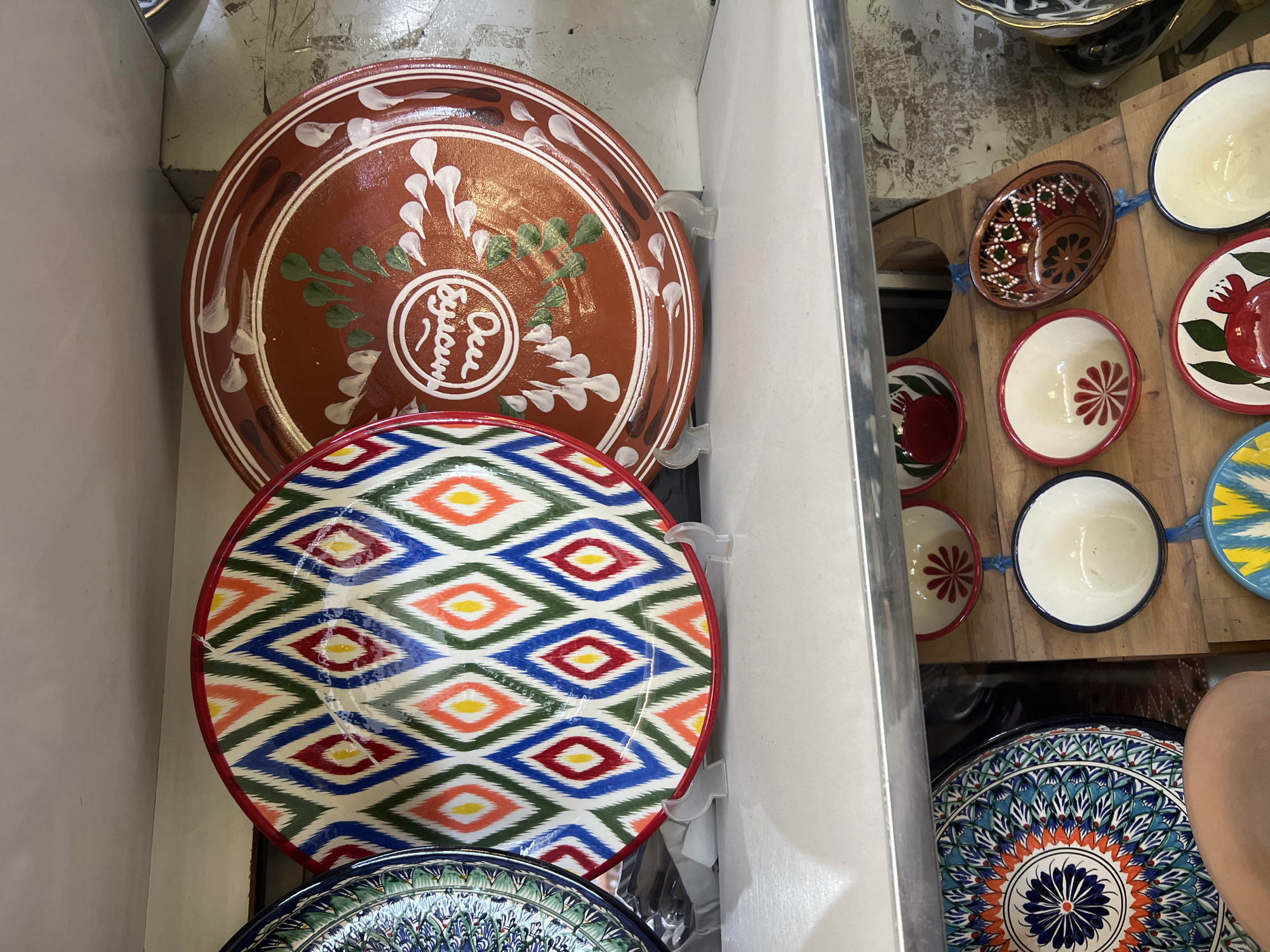 The image depicts a close-up view of a shelf displaying several decorative plates and bowls. In the foreground, on the top shelf, are two large, circular plates. The plate on the left features a vibrant diamond pattern in red, blue, green, yellow, and orange on a white background, with a red rim. The plate on the right is a reddish-brown ceramic with a white circular emblem containing stylized writing and decorative white and green brushstrokes. Both plates are supported by clear plastic stands.
Below the top shelf, on a wooden surface, are several smaller bowls and decorative plates. These items exhibit various patterns, including floral designs and geometric motifs in red, white, and blue. Some of the bowls are suspended by blue cords. In the lower left of the image, a portion of a large, intricately patterned blue and orange plate is visible, along with a person's leg. The background appears to be a wall with a textured, possibly concrete surface. The lighting suggests an indoor environment, likely a market or shop. The presence of traditional Uzbek patterns on the ceramics suggests the location is in Uzbekistan, specifically Eshanguzar, a city known for its handicrafts.