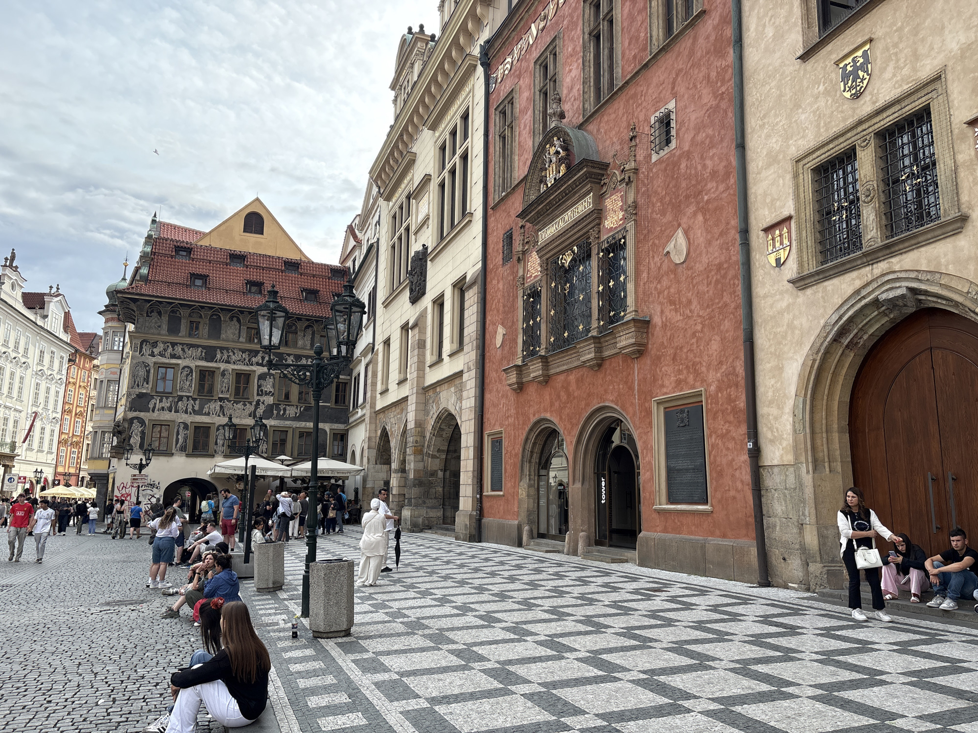 This image captures a view of the Old Town Square in Prague, Czechia. The square features a distinctive checkerboard pattern on the cobblestone ground. On the right, a weathered, red-toned building with arched doorways and windows stands adjacent to a lighter-colored building with ornate architectural details and coats of arms. Several people are visible in the scene: a woman in a white dress holding a black umbrella stands near a lamppost, a group of young people are seated on the curb in the foreground, and other individuals are scattered throughout the square, some walking and others gathered near outdoor seating areas. The sky is overcast, suggesting a cloudy day. The overall impression is of a lively public space with historical architecture.