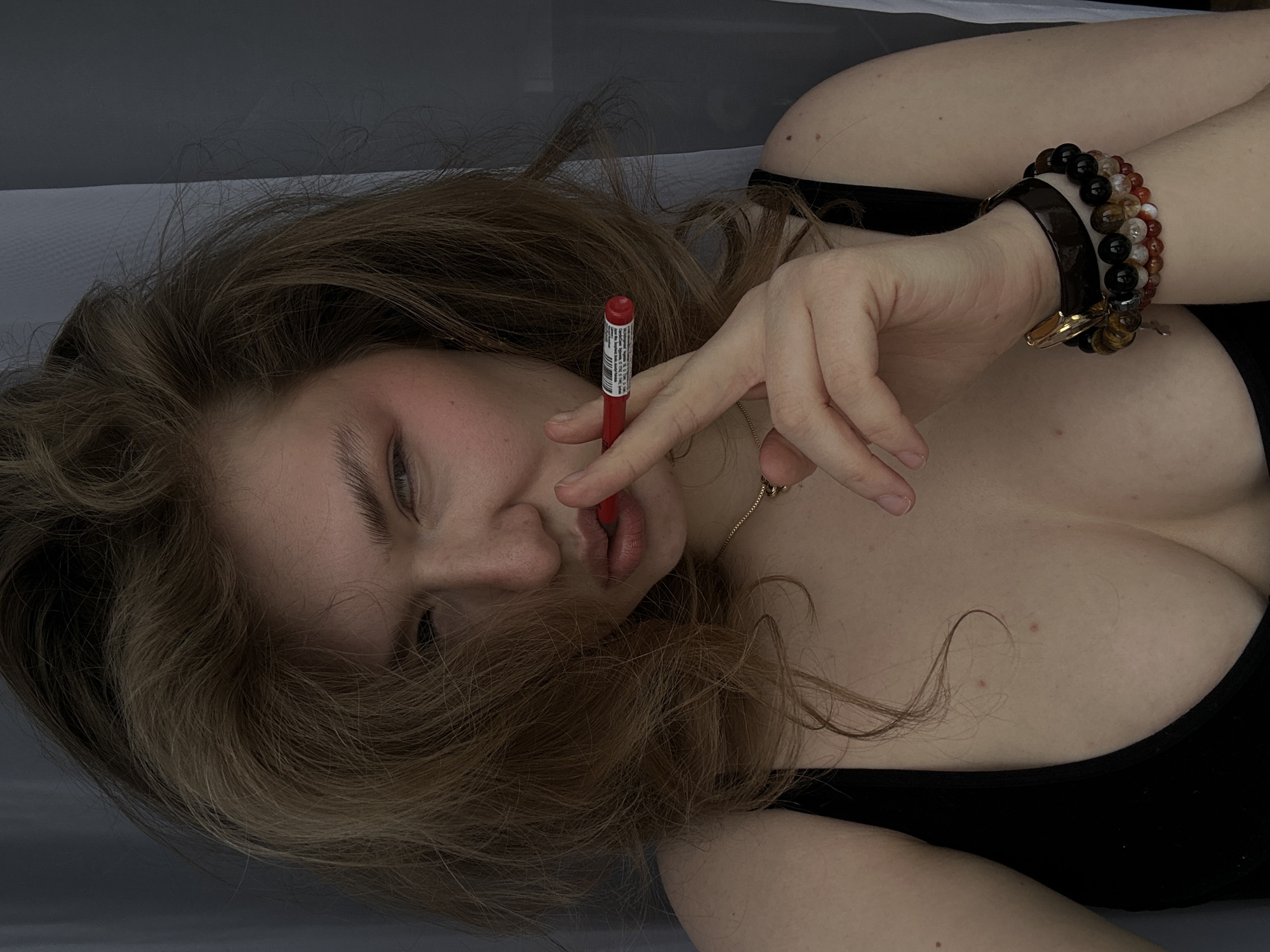 A woman is lying down indoors. She has fair skin, light brown hair, and is wearing a black top. She is holding a red lipstick tube near her lips with her right hand, which has multiple bracelets on the wrist. A gold necklace is visible on her neck. The background is out of focus and appears to be white or light gray. There is no information to identify the location as Nikolskiy, Russia, or any other city.