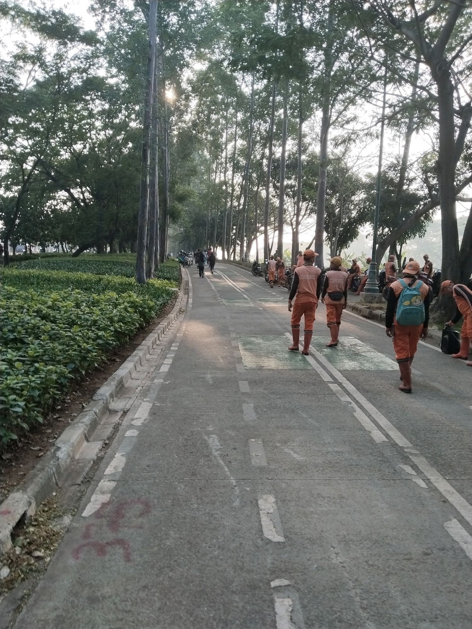 The image depicts a paved path lined with trees and lush green bushes. Several individuals, dressed in orange uniforms, are seen walking along the path. Some are carrying bags, and one is wearing a light blue backpack with a cartoon character. In the background, a few people are riding motorcycles. The scene appears to be a park or a well-maintained roadside area, possibly in East Jakarta, Indonesia, given the context provided. The lighting suggests it is daytime, with sunlight filtering through the trees. The path has white markings, indicating it might be a road or a dedicated walkway.