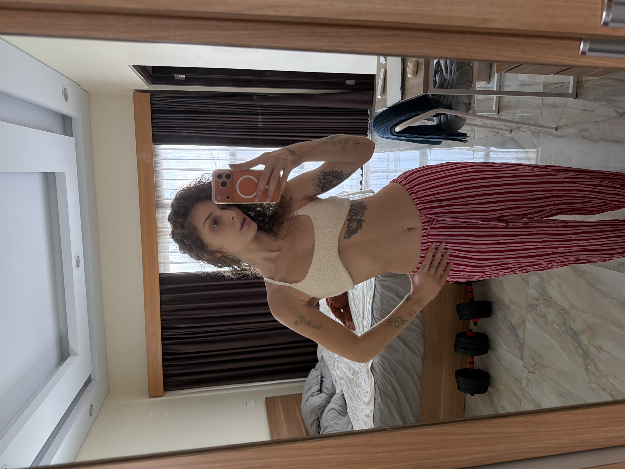 A woman is taking a selfie in a bedroom with a mirror. She is wearing a white textured bralette and red and pink striped pajama pants. She has tattoos on her arms and torso. She is holding an orange-cased phone in her right hand. The room has a bed with gray bedding, a wooden headboard, and a brown blanket on it. There are two dumbbells on the floor. A desk with a chair and a blue towel is visible in the reflection. Dark brown curtains cover a window. The floor is tiled with a marble pattern. The ceiling has recessed lighting and a tray ceiling design. The location is North Kuta, Indonesia.