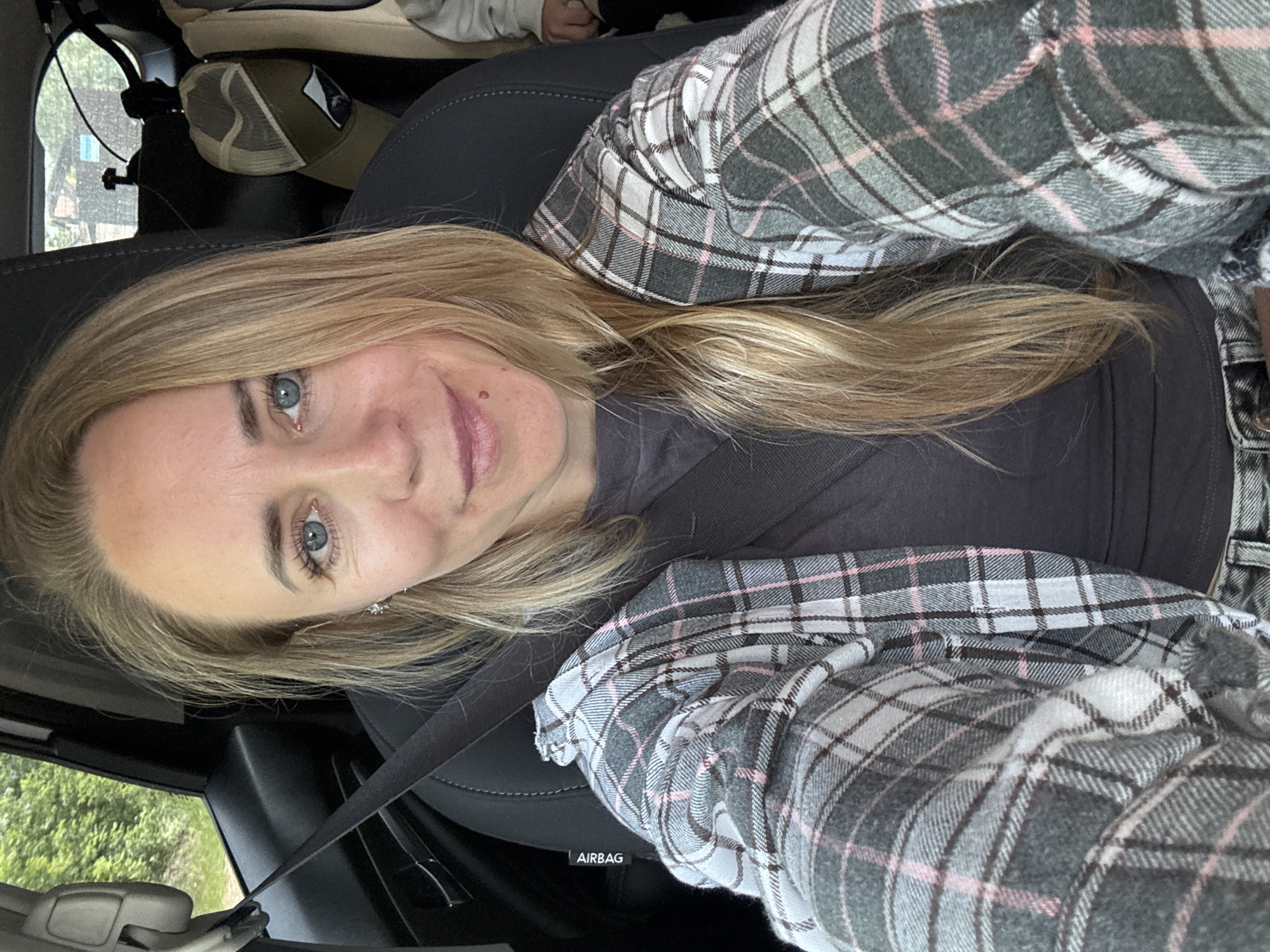 A woman with blonde hair and blue eyes is taking a selfie in the driver's seat of a car. She is wearing a grey and pink plaid shirt over a dark grey t-shirt and a seatbelt. A hat is visible in the background, hanging from the back of the passenger seat. The car appears to be parked on a road surrounded by trees, suggesting an outdoor location.