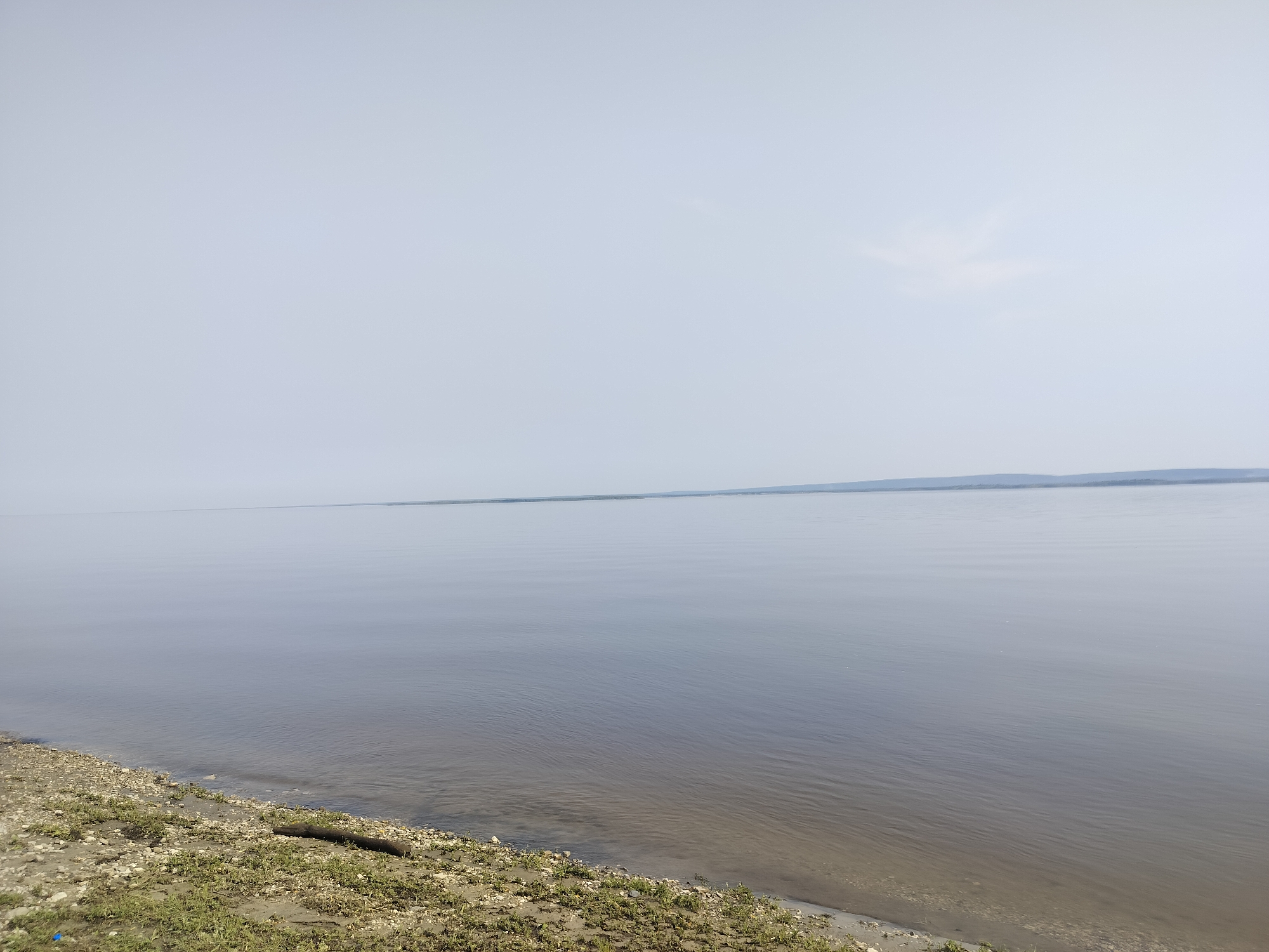 The image depicts a serene, wide body of water, likely a river or a lake, with a calm surface reflecting the overcast sky. The horizon is a faint, almost indistinguishable line where the water meets the distant shore, which appears as a muted, hazy strip of land with a slight elevation. The foreground shows a pebbly and sandy bank sparsely covered with low-lying green vegetation. A dark, cylindrical object, possibly a piece of driftwood, lies partially submerged at the water's edge. The overall atmosphere is tranquil and expansive, characterized by muted colors and a sense of vastness. The scene is consistent with a natural landscape found in the vicinity of Bestyakh, Russia, known for its extensive waterways and natural beauty.