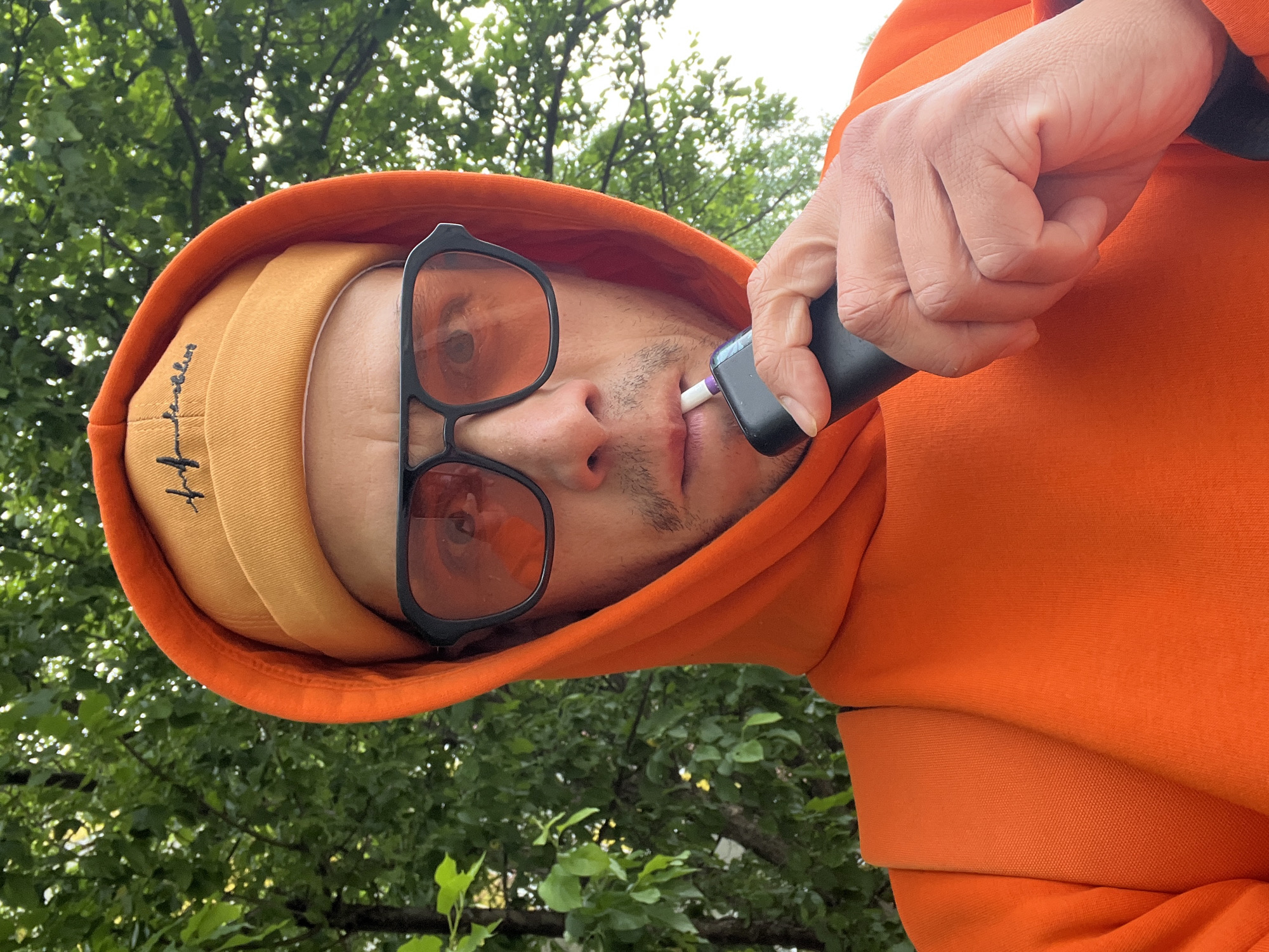 The image is a close-up, low-angle portrait of a person outdoors, rotated 90 degrees clockwise. The subject, who appears to be an adult with light skin and short facial stubble around the mouth and chin, is wearing an orange hooded sweatshirt with the hood up. Beneath the hood, they wear a yellow or tan baseball cap. Dark-framed sunglasses with orange-tinted lenses cover their eyes.
The person is holding a black electronic device, which resembles a heated tobacco product or vape pen, to their mouth with their right hand. A white element with a purple tip is inserted into the device, positioned between their lips. Their right hand, with neatly trimmed fingernails, grips the device.
The background consists of lush green foliage and tree branches, indicating an outdoor setting, possibly a park, garden, or wooded area. The bright, natural lighting suggests it is daytime. The mood is neutral, with the sunglasses obscuring the person's eyes and expressions. On the side of the yellow cap, a small, black embroidered design is visible, appearing as stylized text or a logo that is difficult to decipher definitively, possibly starting with an 'H'. The overall composition frames the person against the natural backdrop, focusing on their face and the device they are using.