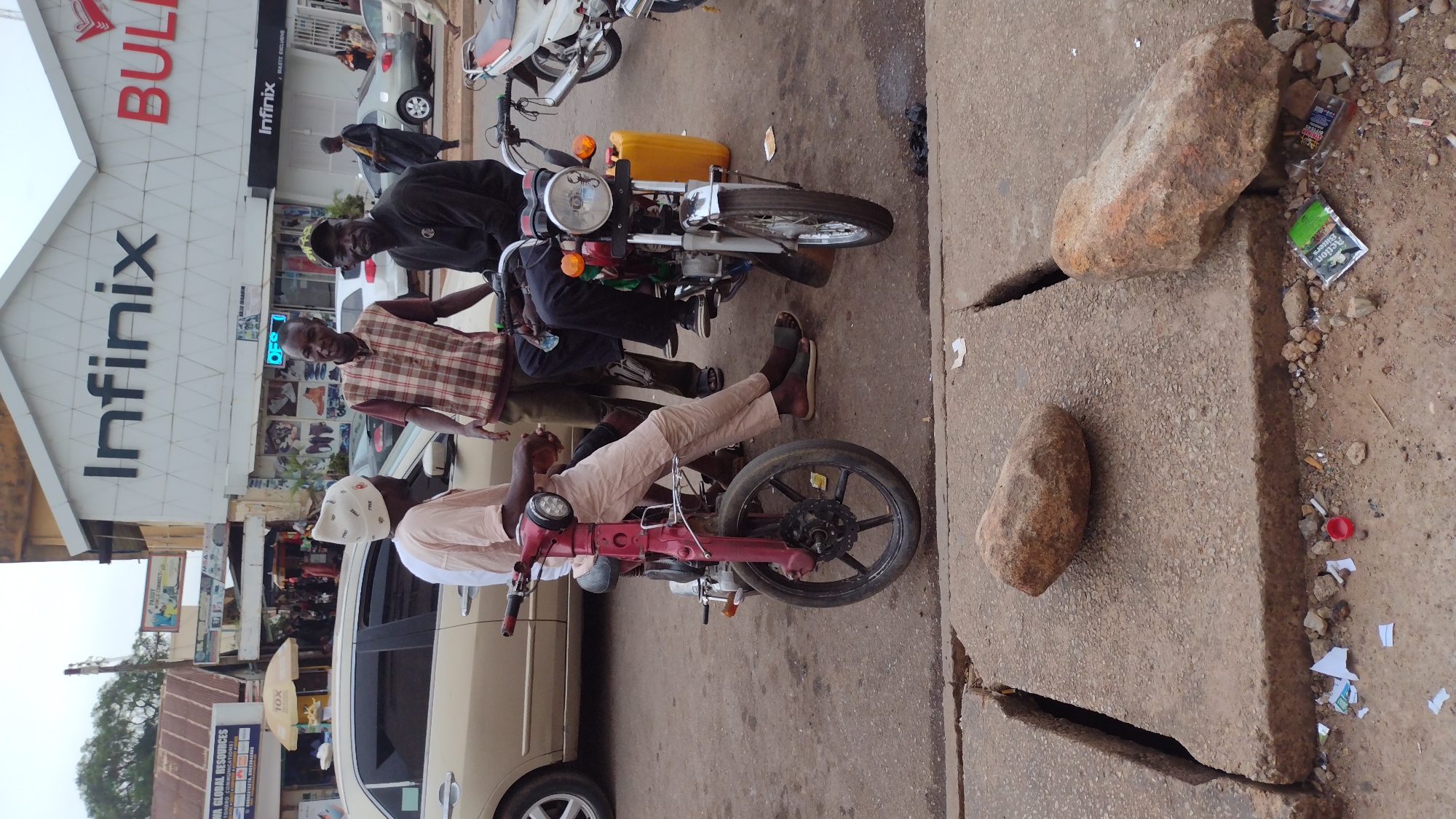 The image shows three men standing next to two motorcycles in front of a building with "Infinix" and "BULL" signage. One man is wearing a pink outfit, a white hat, and riding a red moped style motorcycle. Two men are near a black and silver motorcycle with a yellow gas can strapped to it. One is wearing a plaid vest, and the other is wearing a black shirt. The road is cracked and has rocks placed on it near the curb. Debris is scattered on the ground. A beige car is parked nearby. The location is Jos, Nigeria.