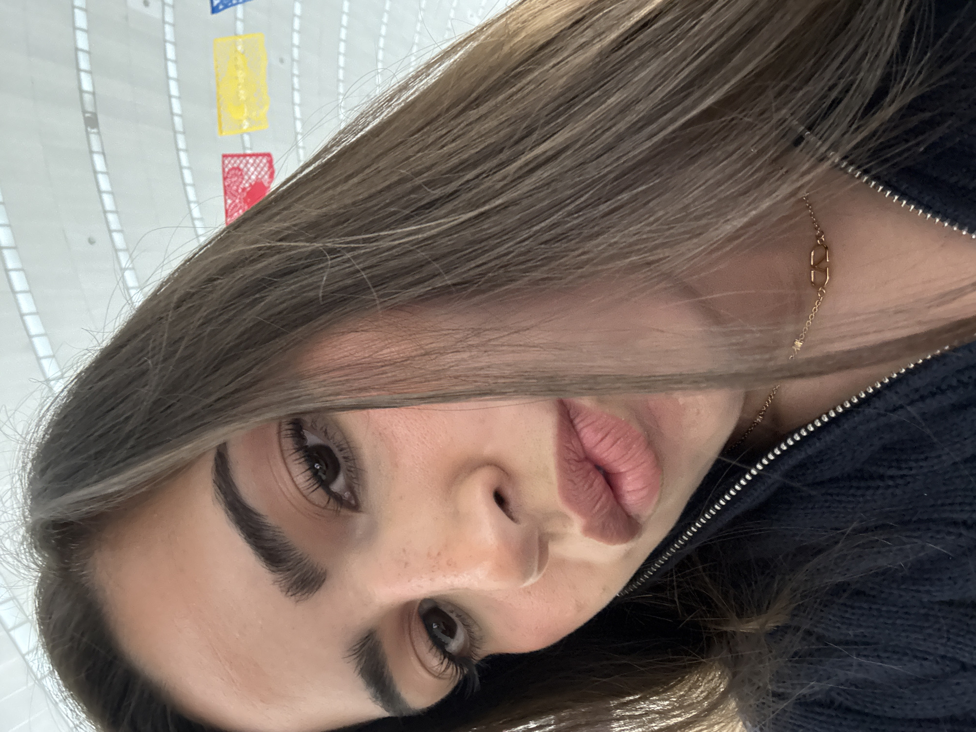 A close-up, angled shot features a woman with light brown hair, visible only from the chest up. She has brown eyes, distinct eyebrows, and pink lips. She is wearing a dark blue zippered jacket, and a gold chain necklace is visible at her neck. The background shows a white wall with colorful rectangular decorations in red, yellow, and blue. The location is San Antonio, United States of America.