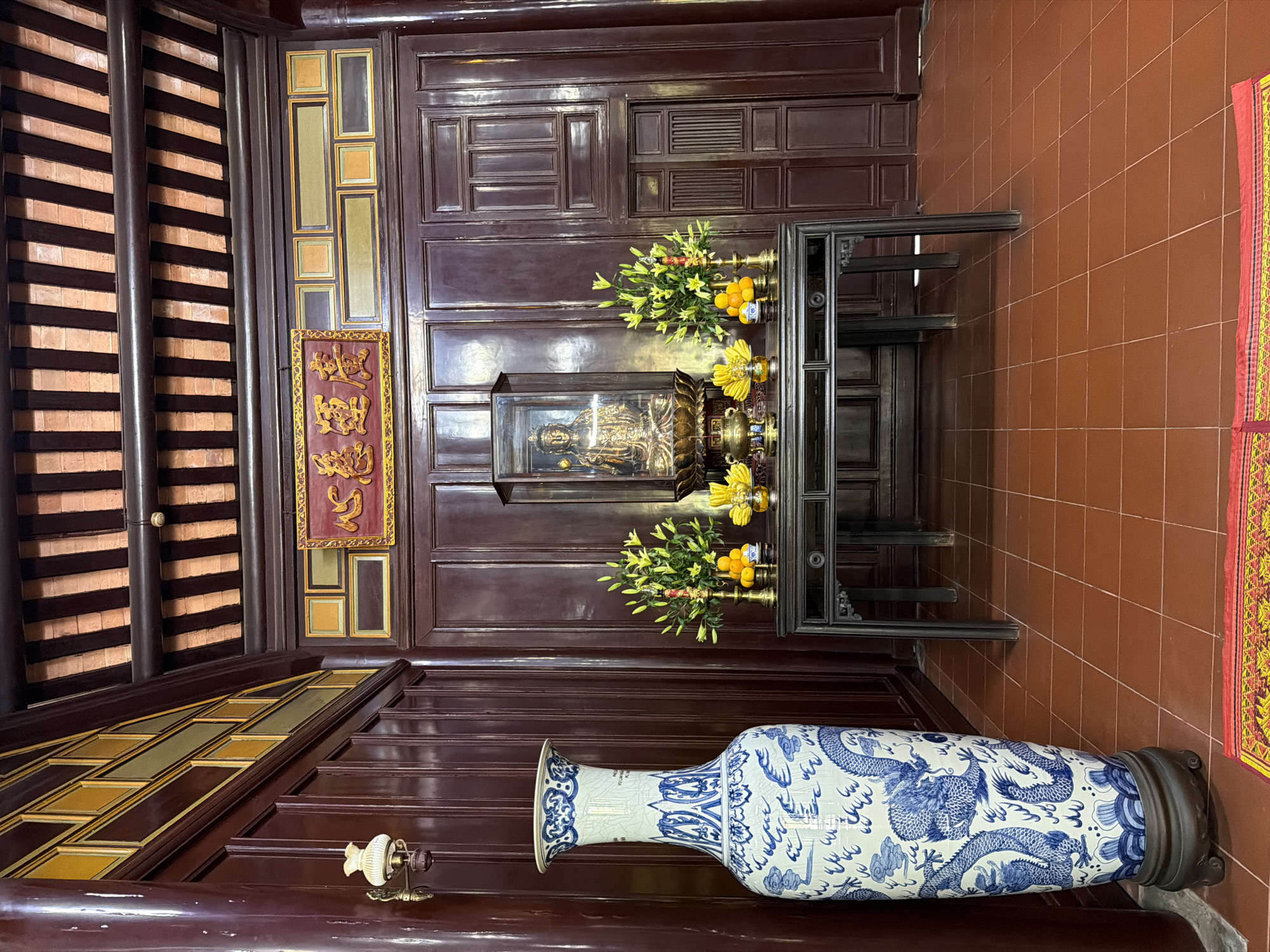 The image depicts the interior of a temple in Kim Long Ward, Vietnam. The scene is dominated by dark wooden panels on the walls and a coffered ceiling with exposed beams. In the center of the room, a dark wooden table displays offerings, including fruit and flowers, in front of a glass case containing a golden Buddha statue. To the left of the table, a large blue and white porcelain vase with dragon motifs stands on a dark wooden base. A traditional lamp is mounted on the wall to the left of the vase. The floor is tiled with reddish-brown square tiles, and a colorful rug is partially visible in the foreground. A sign with golden characters on a red background is mounted on the upper wall.