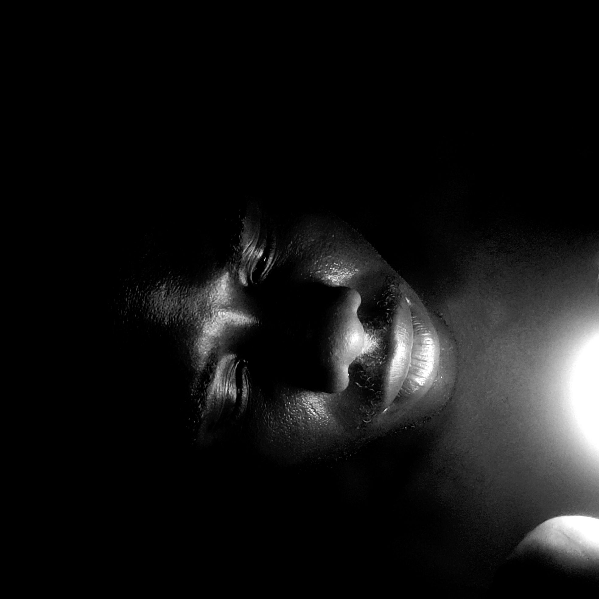 A close-up, black and white portrait captures the face of an individual with dark skin, bathed in dramatic, high-contrast lighting. The person's head is tilted or they are lying down, with their eyes closed or cast downwards, suggesting a state of rest, contemplation, or introspection. A strong, bright light source originates from the right side of the frame, illuminating the profile and contours of the face, including the nose, lips, part of the cheek, and forehead. The skin shows texture and a slight sheen, possibly from moisture or natural oils, particularly on the forehead and around the mouth. The rest of the scene is plunged into deep shadow, rendering the background completely black and emphasizing the subject. A bright, circular light anomaly is visible on the far right, hinting at the source of illumination, possibly a phone flashlight or a small lamp, with a hand partially visible holding it. The image conveys an intimate and serene moment, captured in a very dark setting, likely at night or in an enclosed space. There is no visible text. The location context is Narayi, Nigeria.