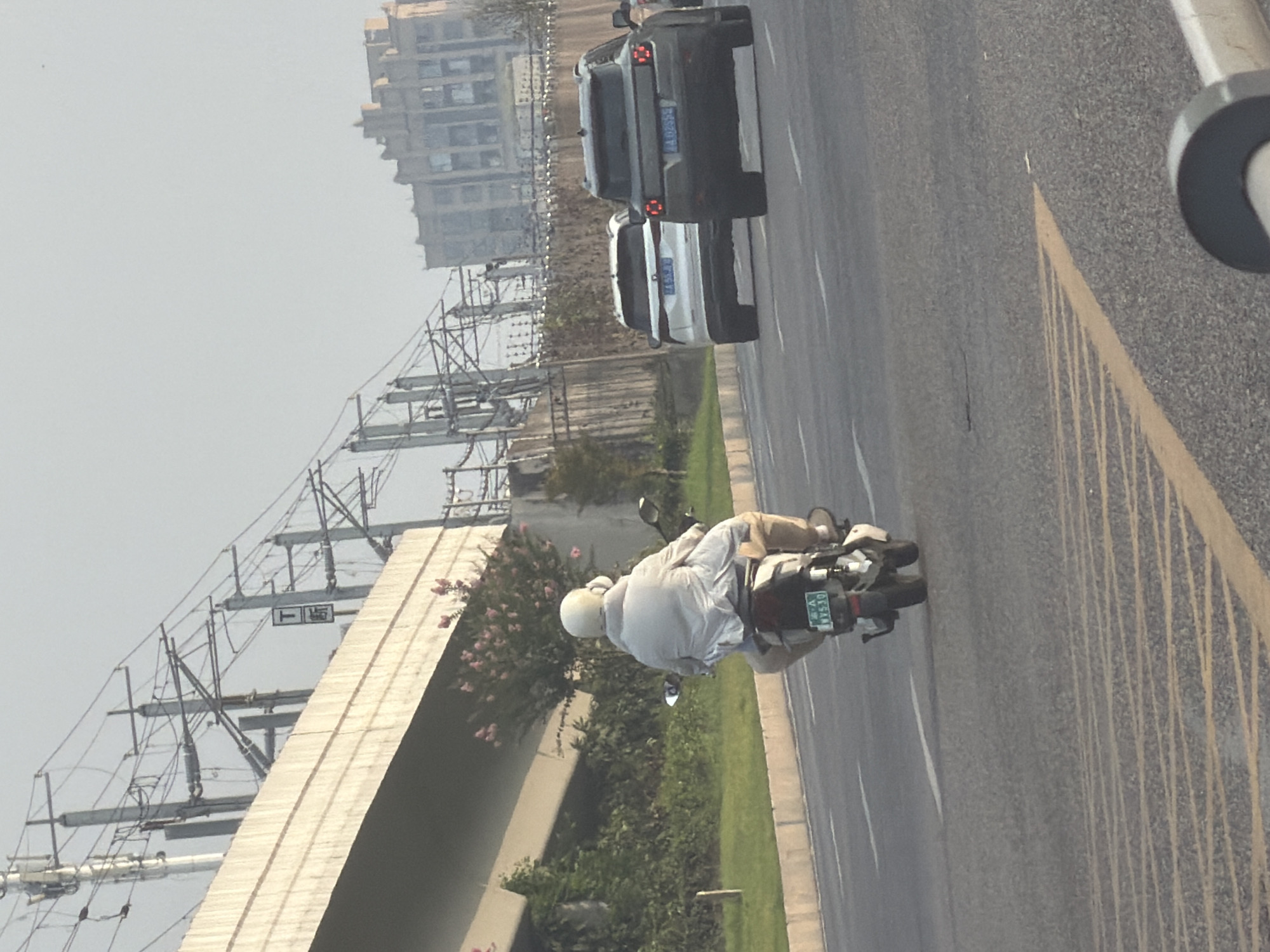 In a city street scene, a person rides a white scooter. The rider, wearing a white helmet and a light-colored puffy jacket, is positioned in the foreground of the image, facing away from the camera. The scooter has a green license plate with the characters "WY530". In the background, two white SUVs are driving in the same direction as the scooter. One SUV has a license plate that is partially visible, reading "JA 55 JIU". Further in the distance, a multi-story building with a distinctive facade is visible. The scene appears to be set in a city environment with overhead power lines and a concrete overpass in the background. The lighting suggests it is daytime, possibly late afternoon due to the soft light. The road is paved and marked with white and yellow lines.