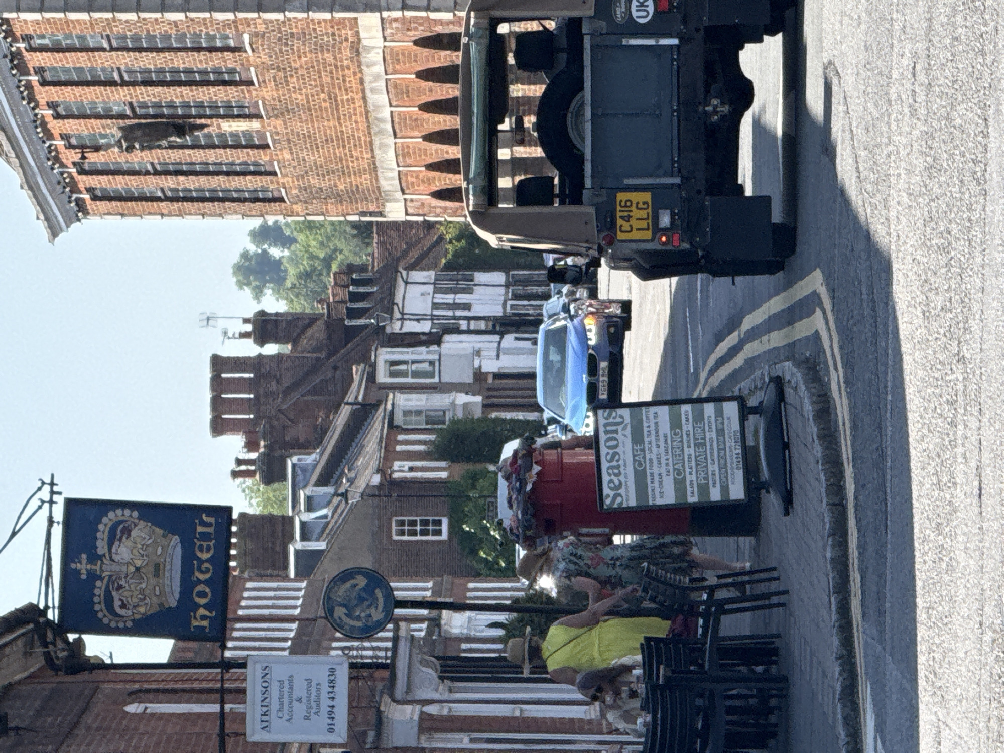 The image captures a street scene in Amersham, United Kingdom, featuring several elements. In the foreground, there's an outdoor seating area belonging to "Seasons Cafe," where people are seated. Adjacent to the cafe is a red postbox decorated with festive garlands. Above the street scene, there's a Hotel sign hanging over a building. A sign with the name of local Chartered Accountants "ATKINSONS" is visible. The street itself has yellow line markings. Several vehicles are present on the road, including a blue car and a tan-colored Land Rover Defender. The Defender's license plate reads "C416 LLG". Buildings with varied architectural styles line both sides of the street.