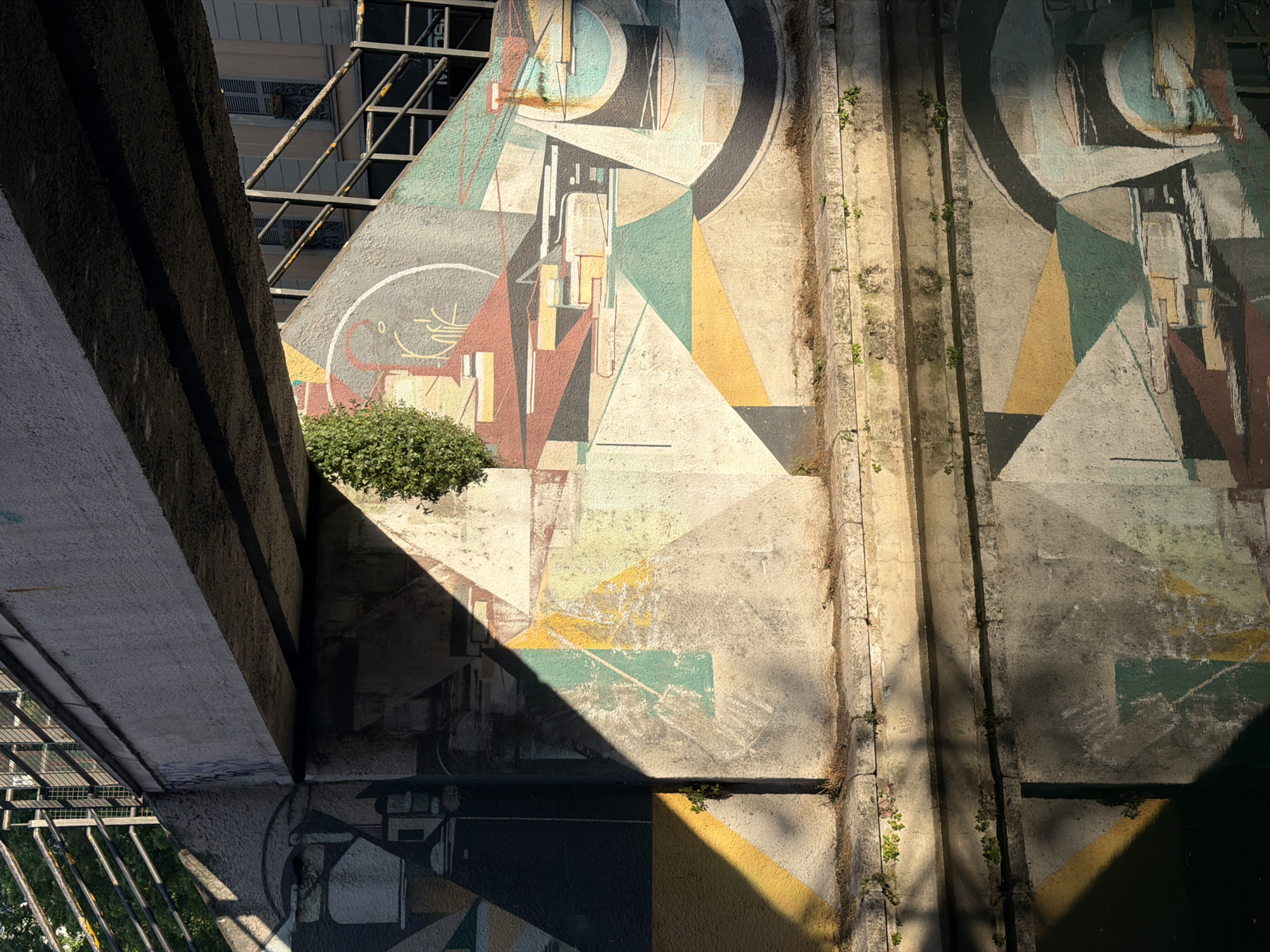 The image depicts an urban scene likely located in Milan, Italy, due to the architectural style and urban infrastructure. A concrete structure, possibly a bridge or an elevated walkway, dominates the foreground. A section of it is painted with a geometric mural in muted colors, including circles, triangles, and rectangular forms. A small shrub grows from the concrete. A section of a metal railing and a building with windows are visible in the background. The scene is bathed in sunlight, which casts sharp shadows across the wall and structure, creating a contrast between light and dark areas. Vertical drainage channels with sparse vegetation run along the concrete surface.