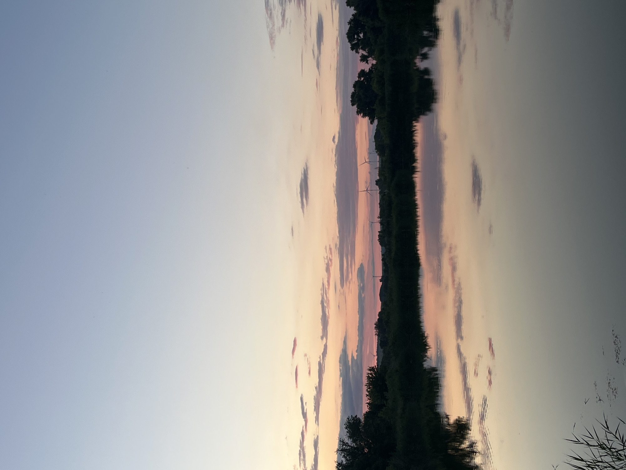 The image depicts a tranquil scene at sunset over a body of water, likely a river or lake, in Kotovo, Russia. The sky is a gradient of pale blue at the top, transitioning through soft pinks and oranges where scattered clouds are visible, reflecting in the calm water below. A dark silhouette of trees and foliage lines the far bank of the water, with several wind turbines visible in the distance on the horizon. The water's surface is smooth, mirroring the sky and the distant landscape. There are no people or animals visible in the frame, and the overall impression is one of peacefulness and natural beauty.