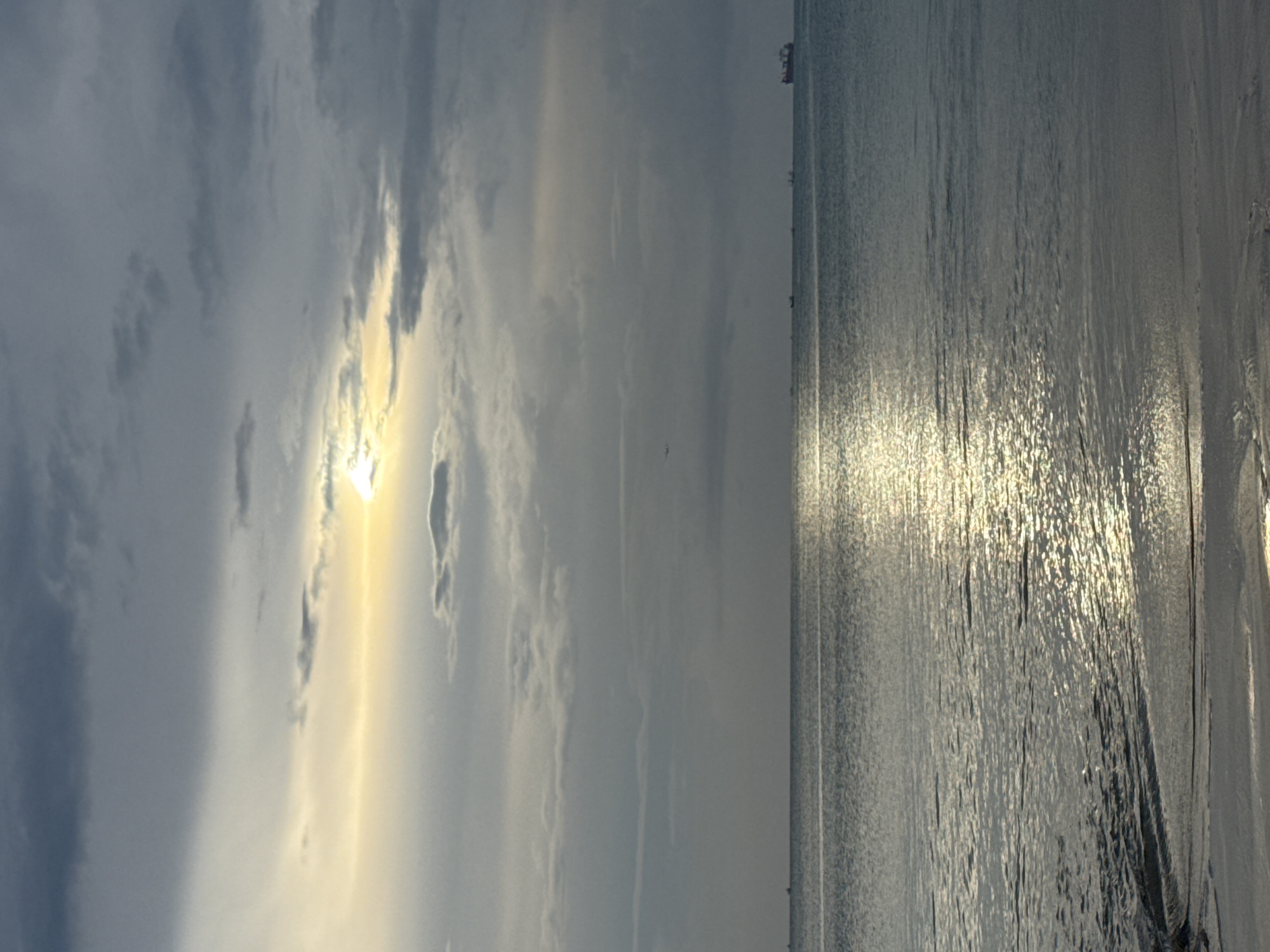 A serene coastal landscape is depicted, likely during either sunrise or sunset. The image shows a vast expanse of water beneath a partially cloudy sky, divided by a distinct horizontal horizon line.
In the upper left portion of the sky, the sun shines brightly, its golden light partially obscured and diffused by layered clouds that vary from dark grey to soft white. Streaks of light pierce through the clouds around the sun.
A prominent, brilliant reflection of the sun stretches across the water's surface, creating a shimmering path that extends from the horizon towards the foreground. The water exhibits a textured appearance with numerous ripples and gentle waves, particularly visible where the sunlight highlights its surface.
Along the distant horizon line, a few very small and indistinct shapes, possibly boats or distant structures, are faintly visible. The overall atmosphere conveys a sense of calm and natural beauty. No people or discernible text are present in the media file. The context for this image is Kuta Selatan, Indonesia.