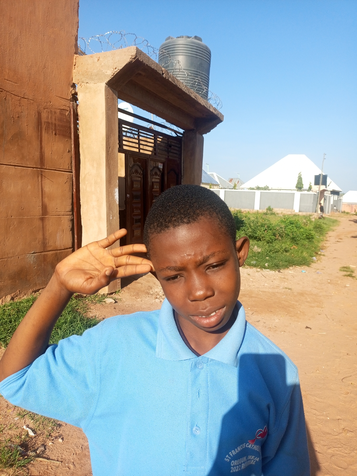 In the city of Tempure, Nigeria, a young boy in a light blue polo shirt stands outdoors in front of a brown building. The boy's left hand is raised to his head, partially obscuring his ear and temple. He looks towards the camera with a serious expression, his mouth slightly open, revealing his teeth. The building behind him has a weathered, textured brown facade and an archway with a metal gate. On top of the archway, a black water tank is visible, surrounded by barbed wire. Beyond the building, a dirt path leads into the distance, flanked by green vegetation and several modern white buildings with angular roofs. The sky is a clear, bright blue. A shadow of the boy's upper body falls across his shirt. The shirt has an embroidered crest on the left breast that reads "ST. FRANCIS CATHOLIC OREGUN, IKEJA 2022 REVIVAL."