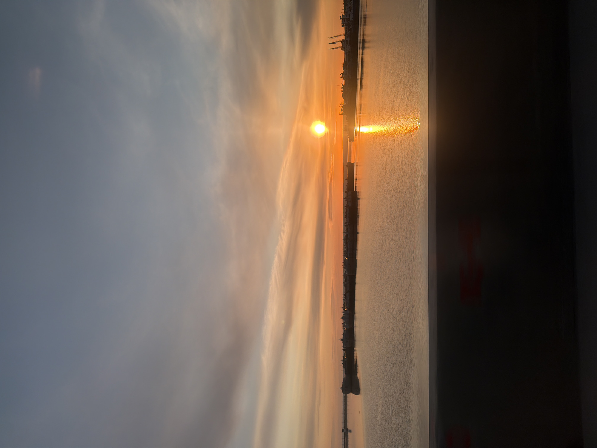 The image shows a sunset over a body of water, possibly a river or bay, in what appears to be Chaikovsky, Russia. The sun is setting on the horizon, casting an orange glow on the water and creating a streak of light reflecting off the surface. A pier or dock stretches out into the water, dotted with silhouettes of structures and possibly industrial buildings. The sky is a mix of gray and orange hues, with wispy clouds. A portion of a wall or structure is visible on the right edge of the frame.