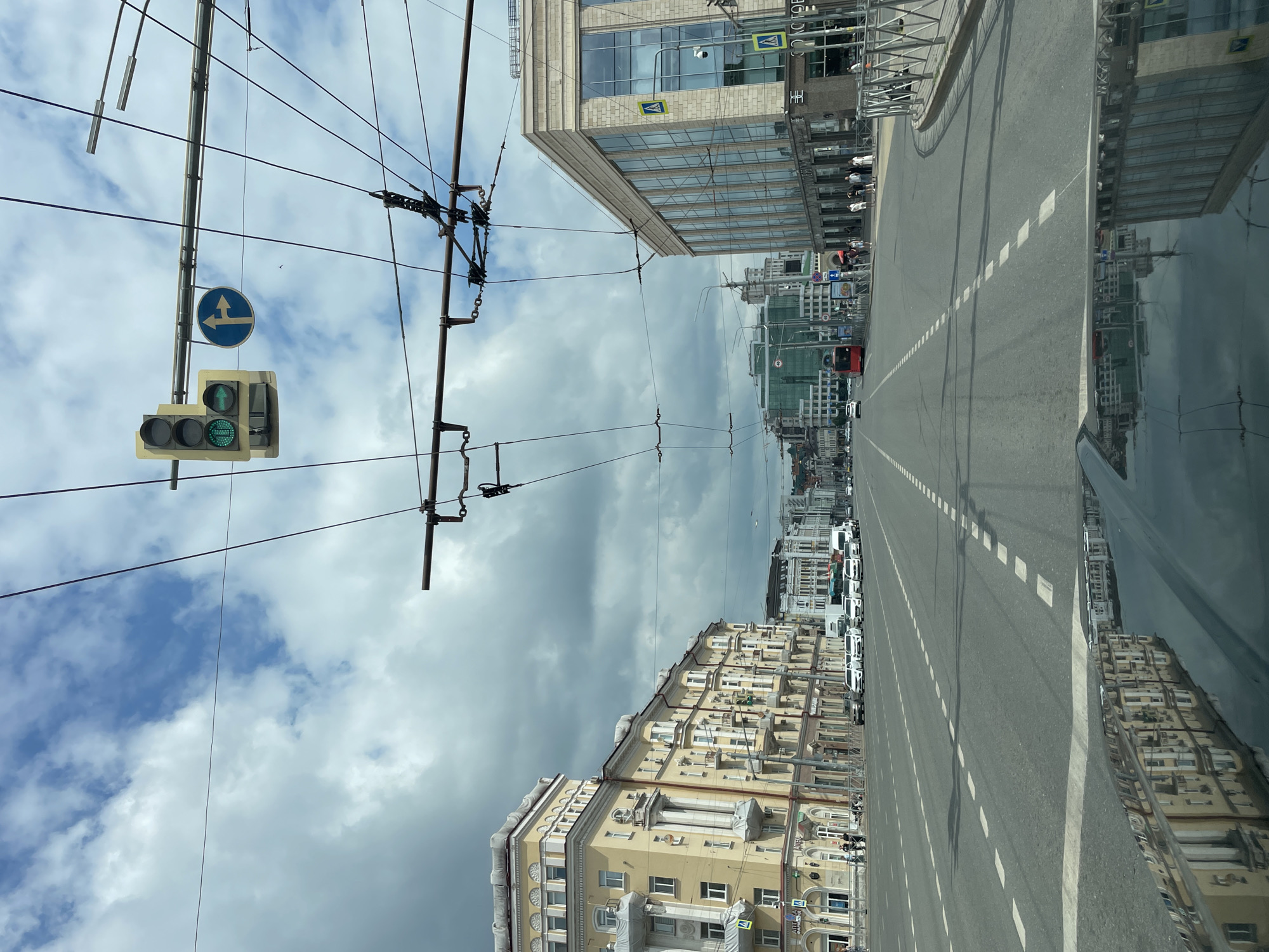The image depicts a street scene in Kazan, Russia. In the foreground, the hood of a dark-colored vehicle is visible, reflecting the buildings and sky. The street itself is wide with multiple lanes and white dashed lane markings. Overhead, numerous electrical wires for trolleybuses crisscross the sky, along with a traffic light displaying a green light for straight ahead and a green arrow for turning right, and a blue circular traffic sign with white arrows indicating a right turn and a straight path.
On the left side of the street, a grand yellow building with ornate white detailing and columns stands prominently. Several other buildings with a similar architectural style line the street. On the right side, a modern, multi-story building with a glass facade and stone detailing is visible. Pedestrians are seen walking on the sidewalks along both sides of the street. Several cars, including white sedans and a red bus, are driving on the road. The sky is partly cloudy with patches of blue sky peeking through. The overall impression is of a busy urban environment.