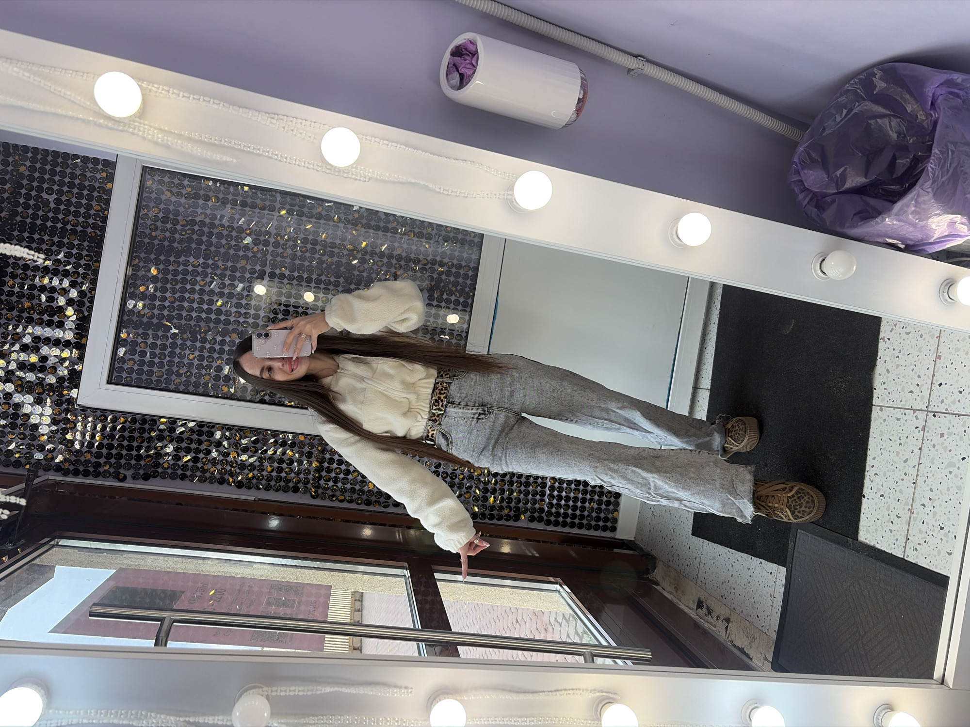 A young woman is taking a mirror selfie in what appears to be a room with a sequined wall. The woman is wearing a cream-colored cropped jacket, light-wash jeans with a leopard-print belt, and leopard-print sneakers. She is holding a cell phone up to her face and smiling.
The mirror is surrounded by a white frame with light bulbs. Outside of the window reflected in the mirror, there are buildings, which might point to an urban setting. Based on the architecture visible in the reflection, specifically the style of the building's exterior and window frames, it is possible the location is in Saint Petersburg, Russia.
The walls of the room appear to be painted in a light purple color. A white object, possibly a hand dryer or some type of dispenser, is mounted to the wall. There is a large bag filled with what appears to be purple fabric or plastic near the top right corner of the mirror.