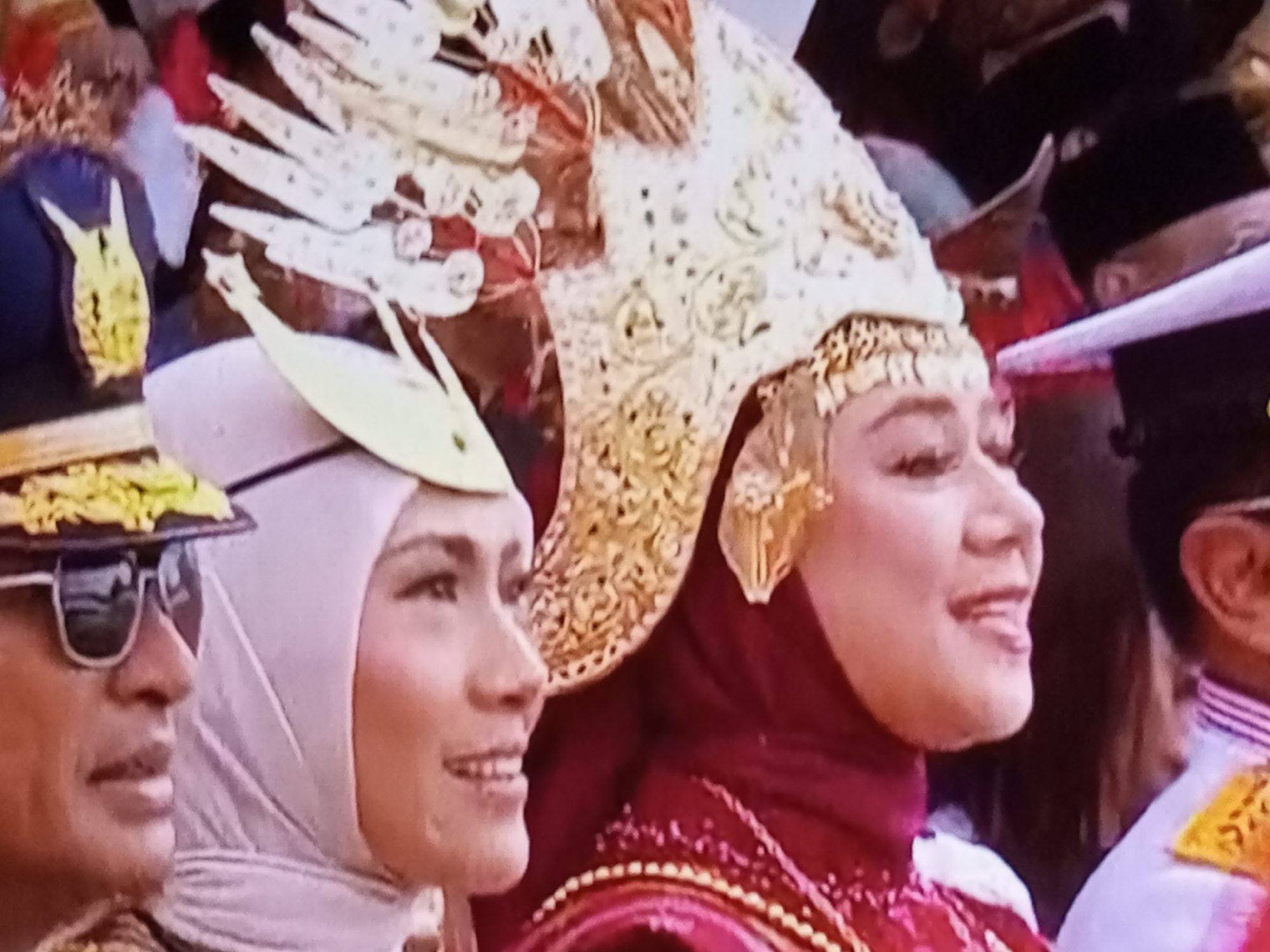 The image depicts a close-up of three individuals in what appears to be a cultural or ceremonial event. The person on the left is a man wearing sunglasses and a hat adorned with gold trim. To his right is a woman wearing a light-colored hijab and a white headpiece. She is smiling and looking to her left. The person furthest right is a woman wearing a red hijab and an elaborate gold headdress that features ornate, feather-like designs. Her head is turned to her left, and she also appears to be smiling. The background is somewhat blurred but suggests a crowd of people. The overall impression is one of celebration or a formal gathering, likely outdoors given the presence of sunglasses. There is no information in the image to confirm a location in Banyudono, Indonesia.