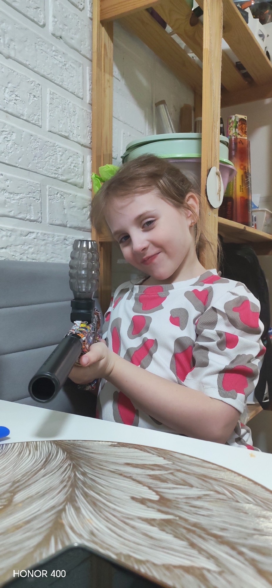 A young female subject is depicted in an indoor residential setting within the city of Noviy, Russia. The subject, appearing to be a child, has light brown hair and is wearing a white short-sleeved shirt patterned with irregular shapes in pink and grey. She holds a toy firearm, characterized by a translucent, ribbed, grenade-shaped magazine, a multicolored body, and a black barrel, aimed slightly downward and to the left. Her head is tilted slightly, and she is smiling directly at the camera.
The background features a white-painted brick wall to the left of the subject. A grey upholstered furniture piece is partially visible behind her left shoulder. To the right, a light wooden shelving unit contains various household items, including a light green bowl, a silver cup, and bottles. The subject is seated at a white table, which has a placemat with a brown and white feather-like design. A small blue object is also partially visible on the table surface. The scene is illuminated by interior lighting.