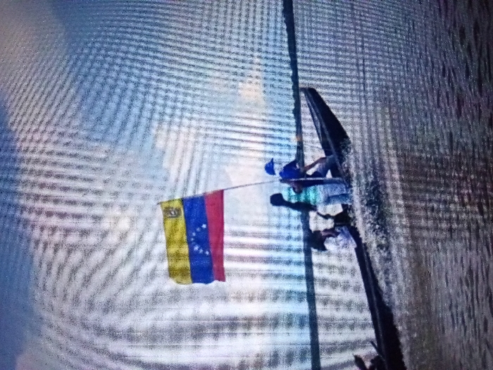 Against a blurry background, a person is shown on a tall structure with a flag of Venezuela flying to the side. The person appears to be working on the structure, possibly doing maintenance or repair. They are wearing a blue hard hat, a light blue shirt, and jeans, and are sitting on a platform attached to the structure. The flag is brightly colored, with yellow, blue, and red stripes and a series of stars in the blue stripe.