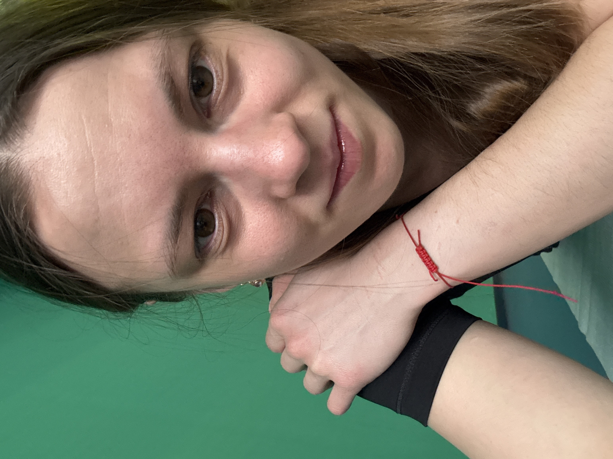 A close-up portrait depicts a young woman with light brown hair and brown eyes, resting her face on her hands. She has fair skin and a slight smile. A red string bracelet is visible on her left wrist. She is wearing a black athletic garment. The background is a uniform green color, suggesting a studio setting or a digitally altered backdrop. The image does not contain enough data to ascertain if it was taken in Chaikovsky, Russia.
