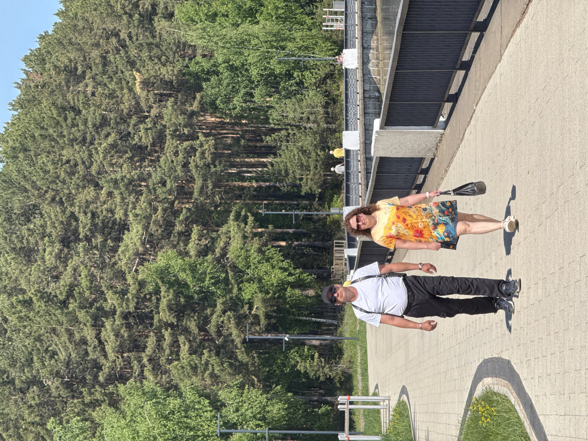 A man and a woman are walking on a sidewalk. The man is wearing a white t-shirt, black pants, and a black baseball cap, and he has a bag slung across his chest. The woman is wearing a yellow dress and sunglasses, and she is carrying a black purse. They are walking on a paved sidewalk next to a building with a black facade. Behind them is a dense forest. The location is Unknown, Unknown.