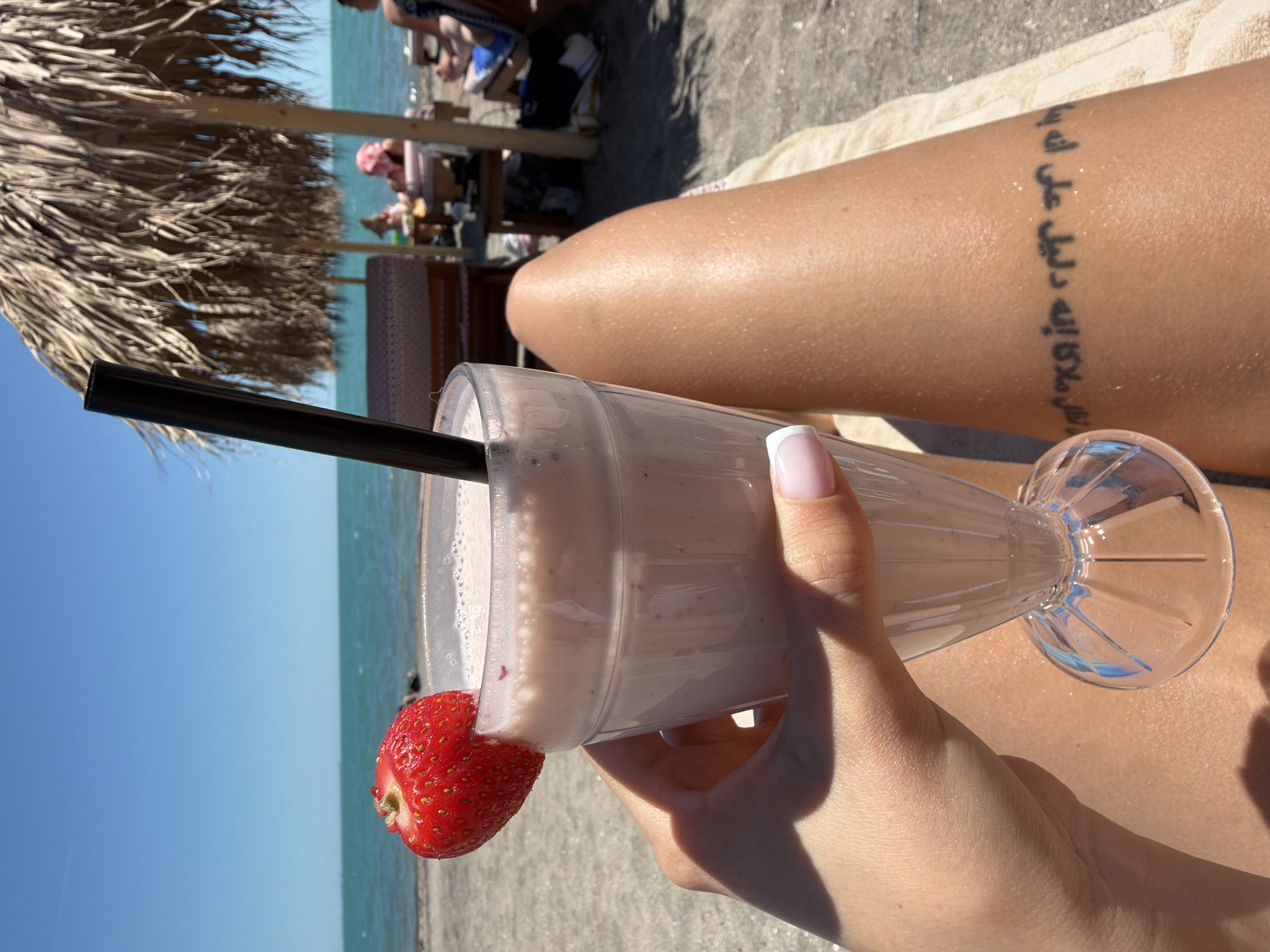 A person's hand, with manicured fingernails, holds a tall, clear glass filled with a light pink smoothie. A fresh strawberry adorns the rim of the glass. A black straw extends from the smoothie. The glass is held against the backdrop of a sandy beach, with the turquoise sea and a clear blue sky visible in the distance. In the background, people relax under a thatched umbrella, some on lounge chairs. The image appears to be taken on a sunny day at a beachside location. The presence of the sea and beach suggests a coastal area, possibly within the city of Burgas, Bulgaria, known for its beaches along the Black Sea coast. A tattoo is visible on the person's leg, featuring what appears to be Arabic script.