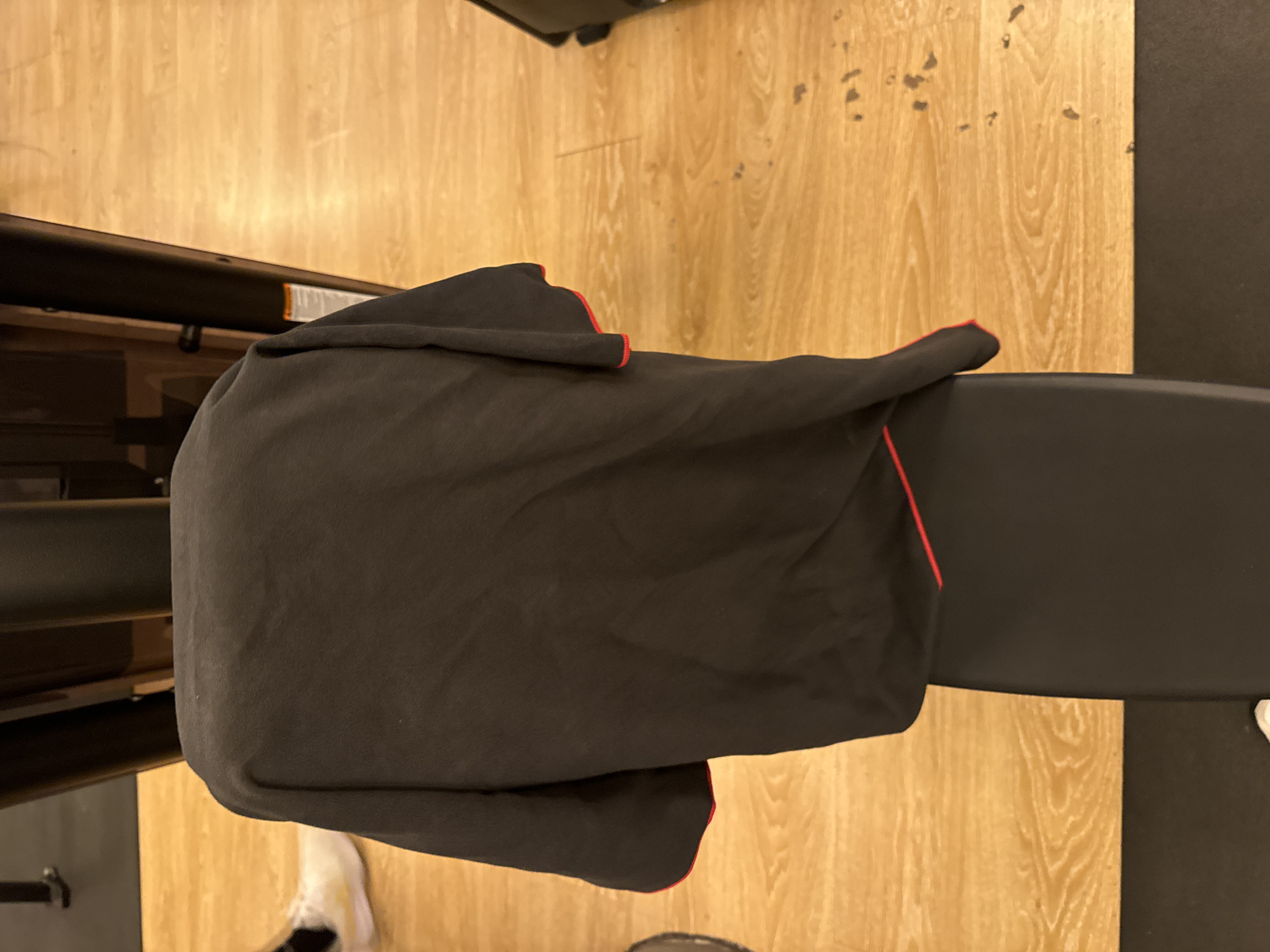 The image shows a black towel with a red trim draped over a black bench. The bench is positioned on a wooden floor. In the background, part of a black exercise machine is visible. A white sneaker is also visible on the floor near the bench. The scene appears to be indoors, likely in a gym or fitness facility, given the presence of exercise equipment and the type of flooring. There are no clear indicators of time of day or weather conditions. No text is clearly legible in the image. The focus of the image is the towel and the bench, suggesting the towel might be for personal use during a workout.