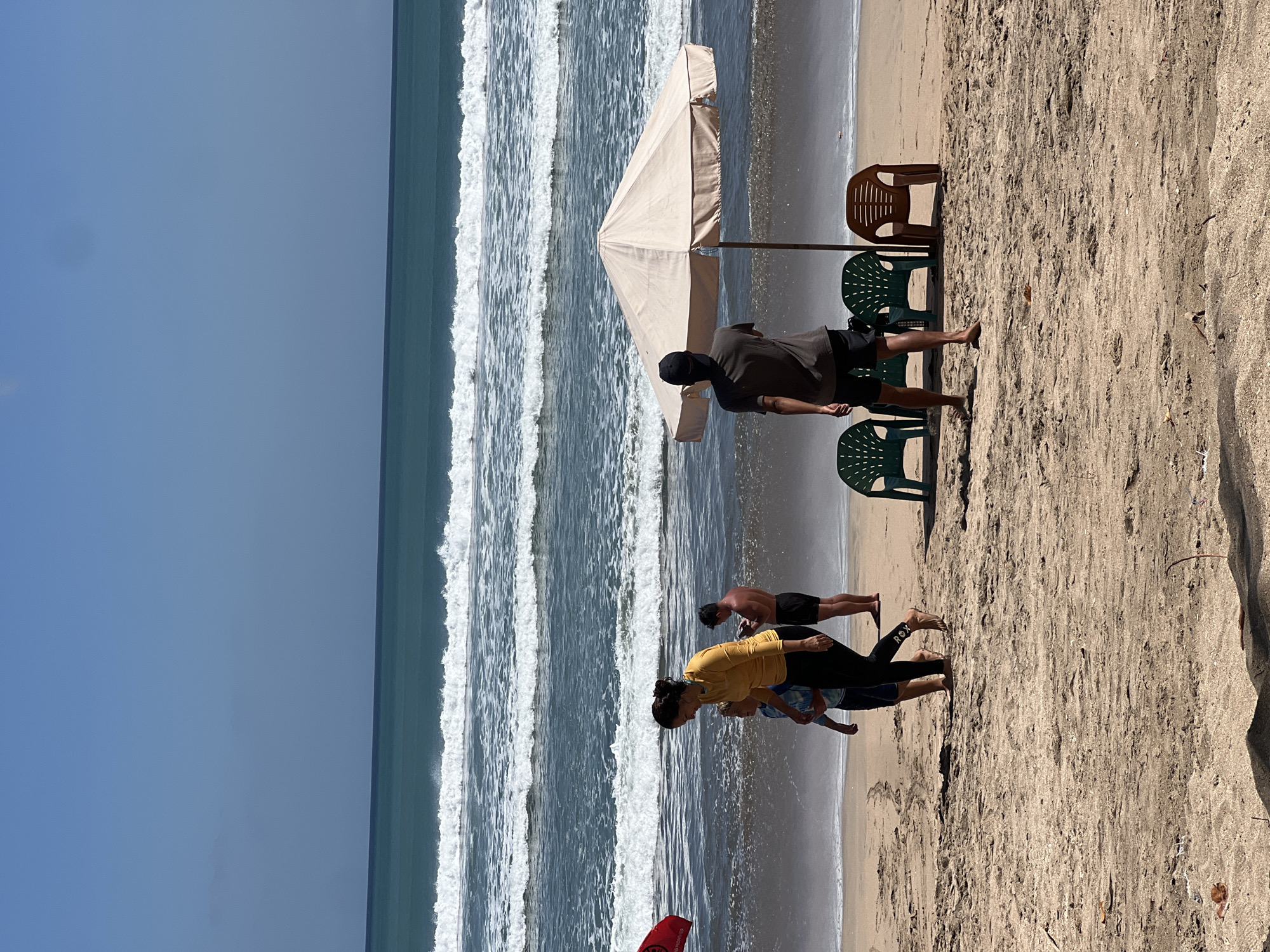 This image captures a sunny day at Kuta Beach in Indonesia. The foreground shows sandy beach with a few scattered leaves and debris. In the middle ground, several people are present. A woman in a yellow top and black leggings, accompanied by a child, walks across the sand. Nearby, a man in black shorts stands. Further to the right, under a large beige beach umbrella, three plastic chairs are arranged: two green ones on the left and a brown one on the right. A person wearing a dark t-shirt and a black cap stands facing the ocean, near the umbrella. In the background, the turquoise ocean features several rows of white-capped waves rolling towards the shore under a clear blue sky. A red flag is visible on the left side of the image.