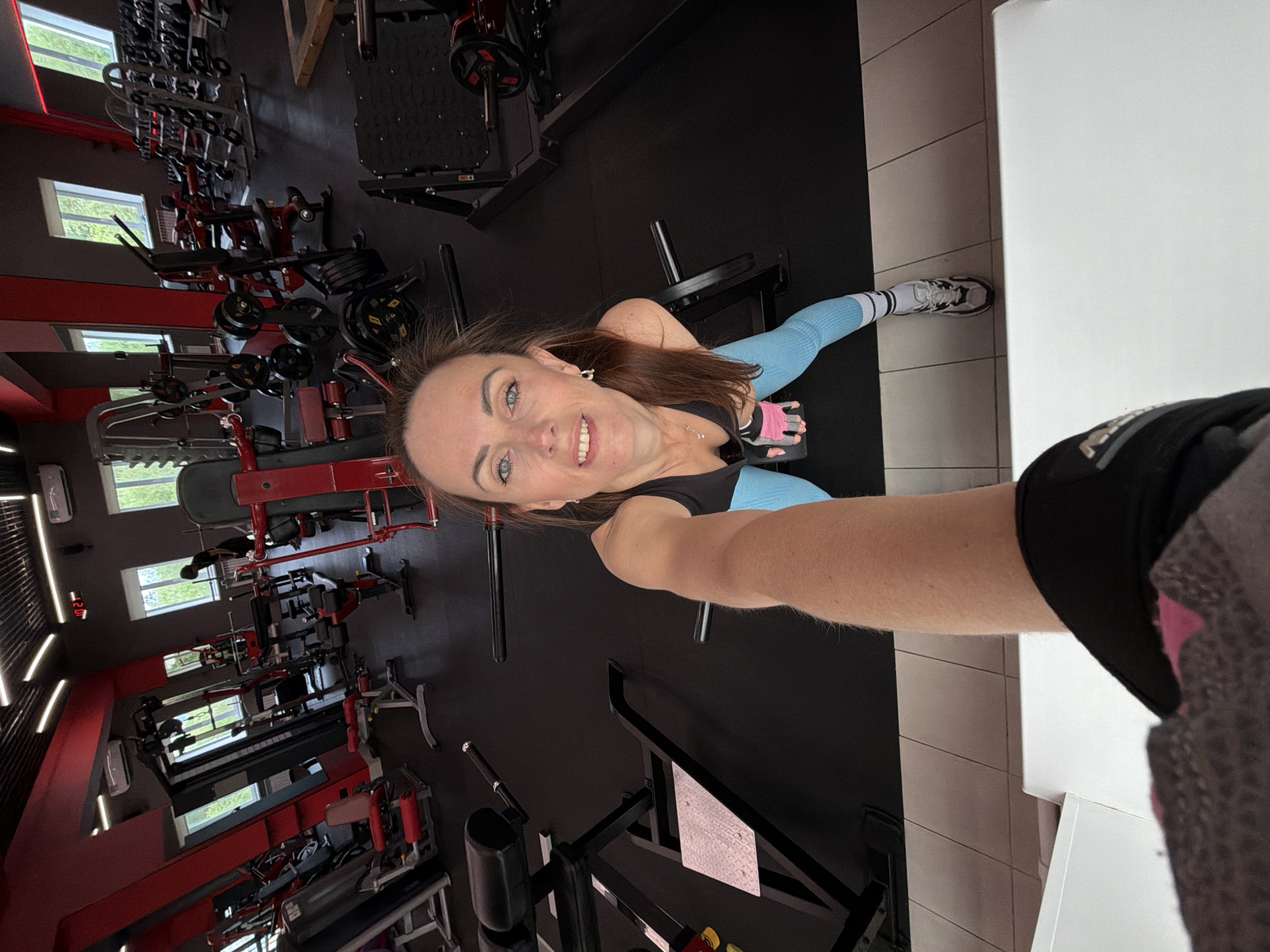 A woman with brown hair and blue eyes is taking a selfie in a gym. She is wearing a black sports bra, light blue leggings, and white athletic shoes. She has pink fingerless gloves on her hands and is wearing a silver necklace. The gym has black flooring and red accents on the equipment. There are various weight machines and free weights visible in the background. The scene appears to be indoors in a well-lit gym.