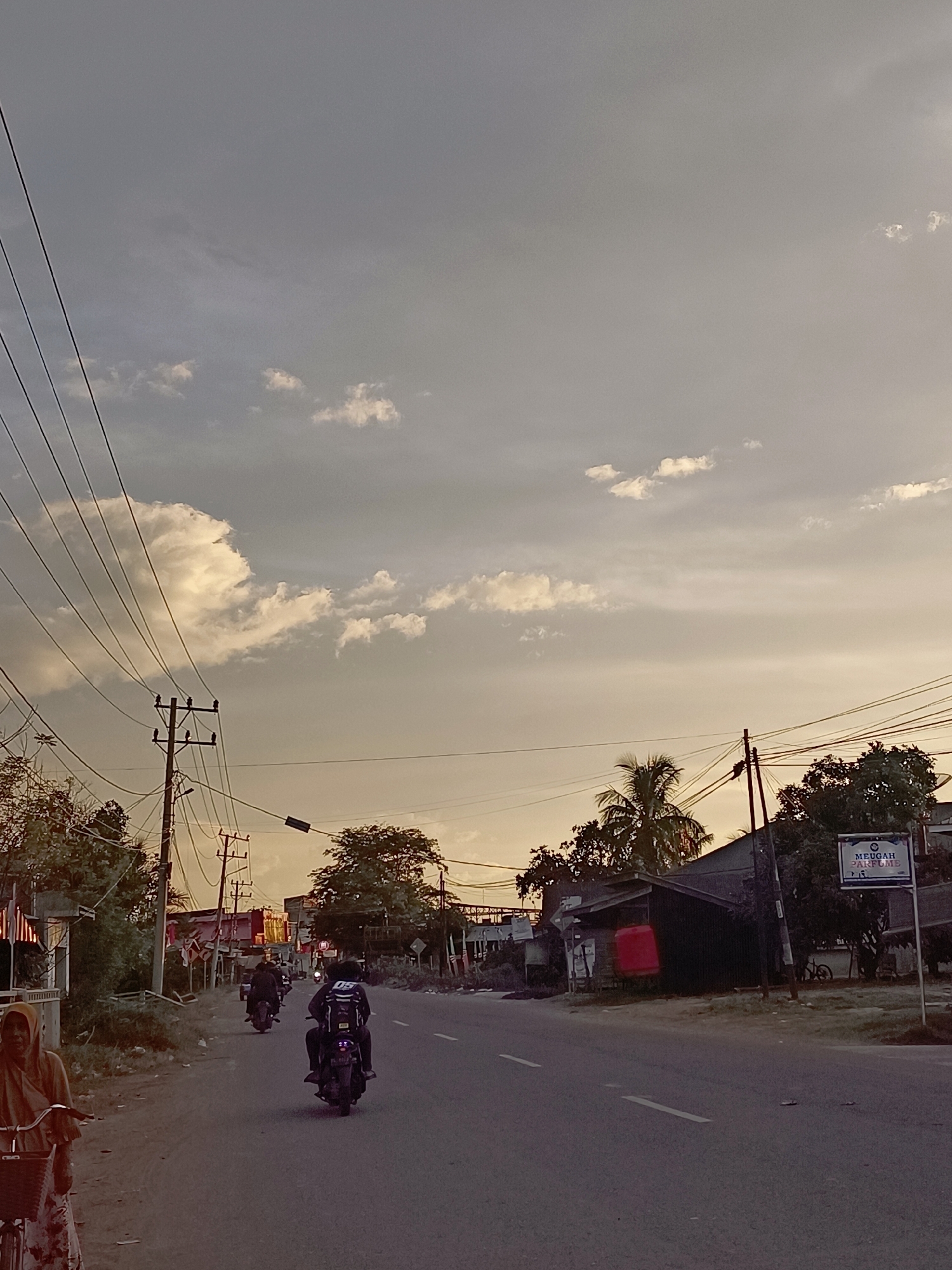 The image depicts a street scene in Blang Cut, Indonesia, during what appears to be late afternoon or early evening. The sky is overcast with diffused sunlight, casting a soft, warm glow. Multiple utility lines crisscross the sky, supported by wooden poles. On the left side of the road, a woman wearing a headscarf and a patterned skirt is partially visible, standing next to a bicycle. Further down the road, several people are riding motorcycles. One motorcycle, in the foreground, is ridden by a person wearing a dark jacket with white lettering and a helmet. Another motorcycle is further ahead, and a third is in the distance. The right side of the road features buildings with dark roofs, some appearing to be shops or homes, and lush green trees. A sign with the text "MEUGAH PARFUME" and a logo is visible near a building. The road itself is asphalt, with white lane markings. The overall atmosphere is calm and rural.