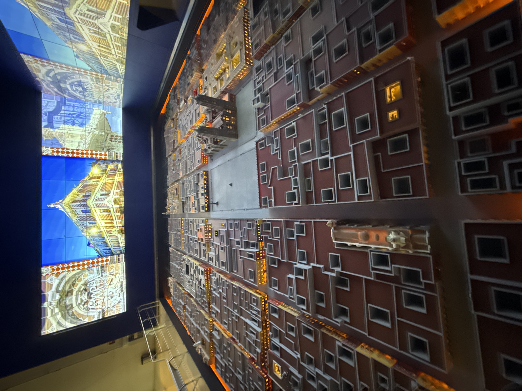 The image shows an art installation. On the left, there are three video screens displaying detailed images of European-style architecture, with blue skies visible. To the right of these screens is a large, intricate model of a city. The model features rows of buildings, mainly in shades of red, brown, and grey, with numerous windows and architectural details. Many of the windows appear to be illuminated with a yellow/orange light. The perspective suggests the installation is viewed from below, looking upward. There is a handrail at the bottom left of the image, suggesting a viewing platform or area.