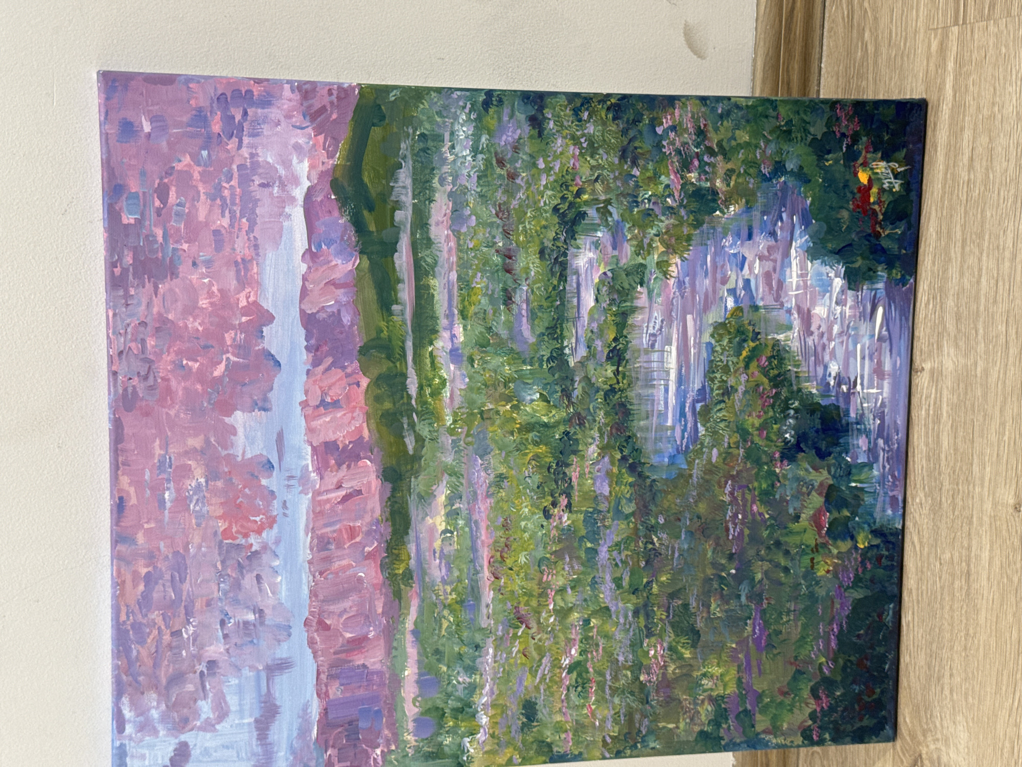 The image shows a painting of a scene resembling a nature-filled landscape. The left portion of the painting features a light blue area bordered by pink and lavender brushstrokes, giving the impression of a waterfall. The rest of the painting is dominated by green and purple hues, which seem to depict vegetation, possibly trees and flowers. In the upper right corner, there are some red and yellow brushstrokes. The painting appears to be resting against a white wall and a wooden panel. There is no information present to associate the scene with the city of Yerevan, Armenia.