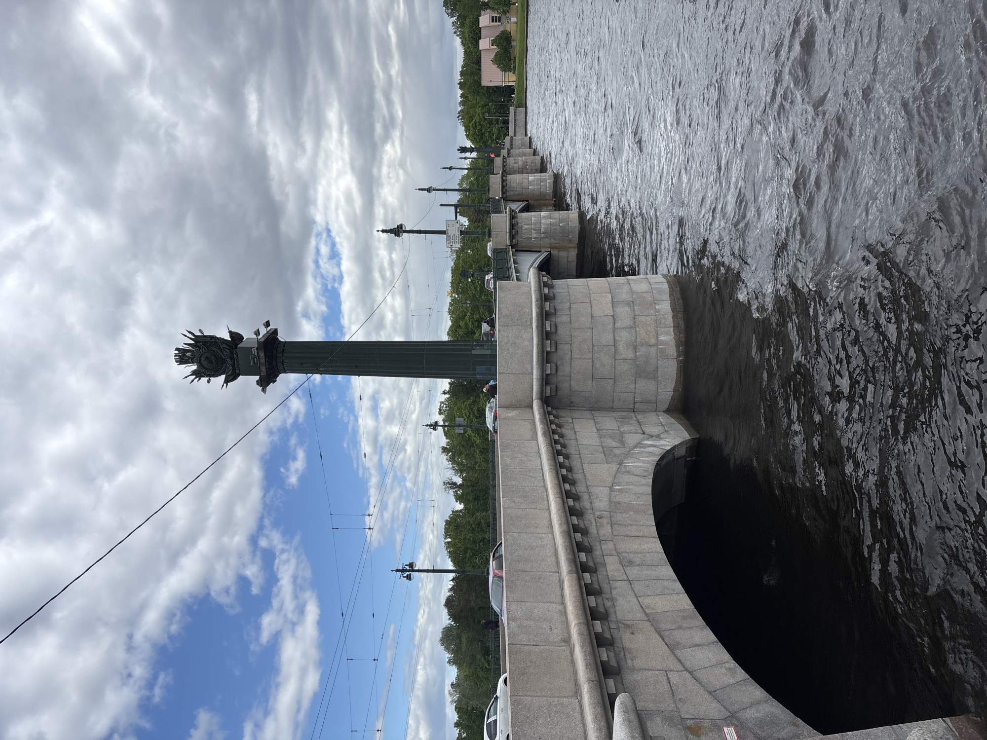 The image shows a view of the Rostral Columns and Liteyny Bridge in Saint Petersburg, Russia. The Rostral Columns are tall, decorative columns topped with ship prows and statues. The Liteyny Bridge is a drawbridge with granite arches and railings. The Neva River flows beneath the bridge. The sky is cloudy with patches of blue. Cars are present on the bridge.