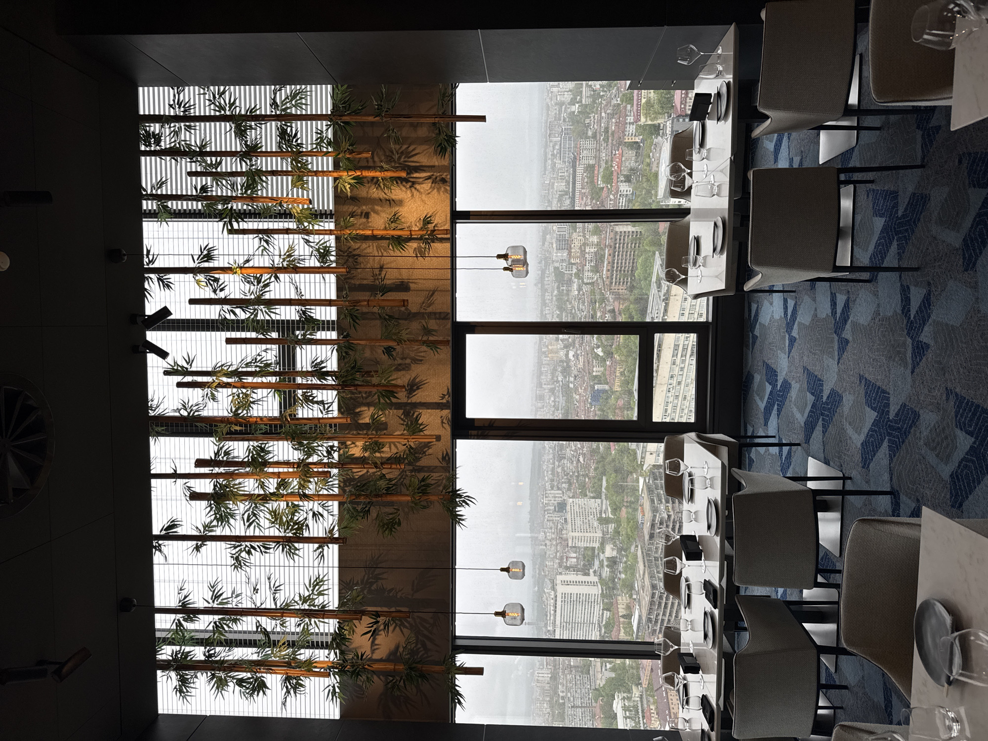 This media file captures the interior of a modern, high-rise restaurant or dining area in Sofia, Bulgaria, offering panoramic views of the city. The space is elegantly designed with a focus on contemporary aesthetics and a connection to nature.
The most striking feature is an elaborate ceiling installation covering a significant portion of the overhead space. This installation comprises parallel wooden beams, resembling bamboo stalks, intertwined with artificial green leaves. Behind these decorative elements, illuminated slatted panels create the effect of a bright, stylized skylight, providing a soft, diffused light. A dark, solid ceiling runs along the left side of the room, where a circular vent is visible.
Along the right side of the room, large, floor-to-ceiling windows present an expansive view of a densely built urban landscape. The sky appears overcast, casting a somewhat muted light over the city, which consists of numerous buildings of varying heights and architectural styles. In front of the windows, two minimalist glass pendant lights with visible filaments hang, adding to the contemporary decor.
The dining area is furnished with several rectangular tables, meticulously set with white placemats or tablecloths, dark and light plates, and an assortment of wine glasses, indicating readiness for service. Modern, armless dining chairs, upholstered in a light grey fabric with dark frames, are arranged around the tables. The floor is covered with a patterned carpet featuring geometric shapes in shades of blue, grey, and white.
No people are visible in the image, suggesting it was taken either before opening hours or during a quiet period. The overall atmosphere conveys a sense of sophisticated tranquility. There is no visible text.