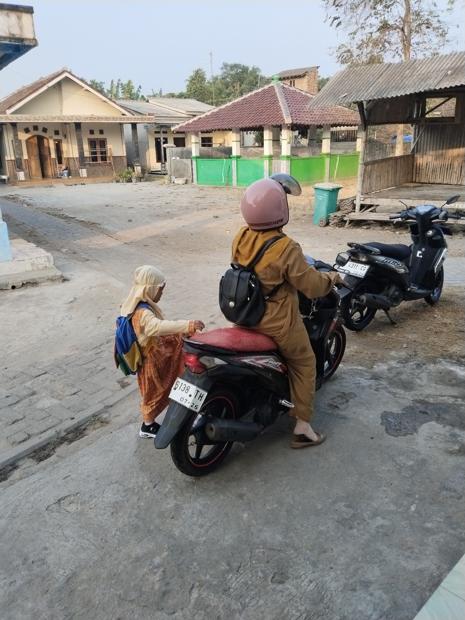 In the city of Serang, Indonesia, two individuals are positioned near motorcycles on a concrete surface. One individual, wearing a tan jumpsuit and a pink helmet, is seated on a black motorcycle. Beside them, a child in a tan outfit and hijab, carrying a colorful backpack, stands with one foot on the rear of a red-seated motorcycle. The background reveals a residential area with houses and a shaded structure.