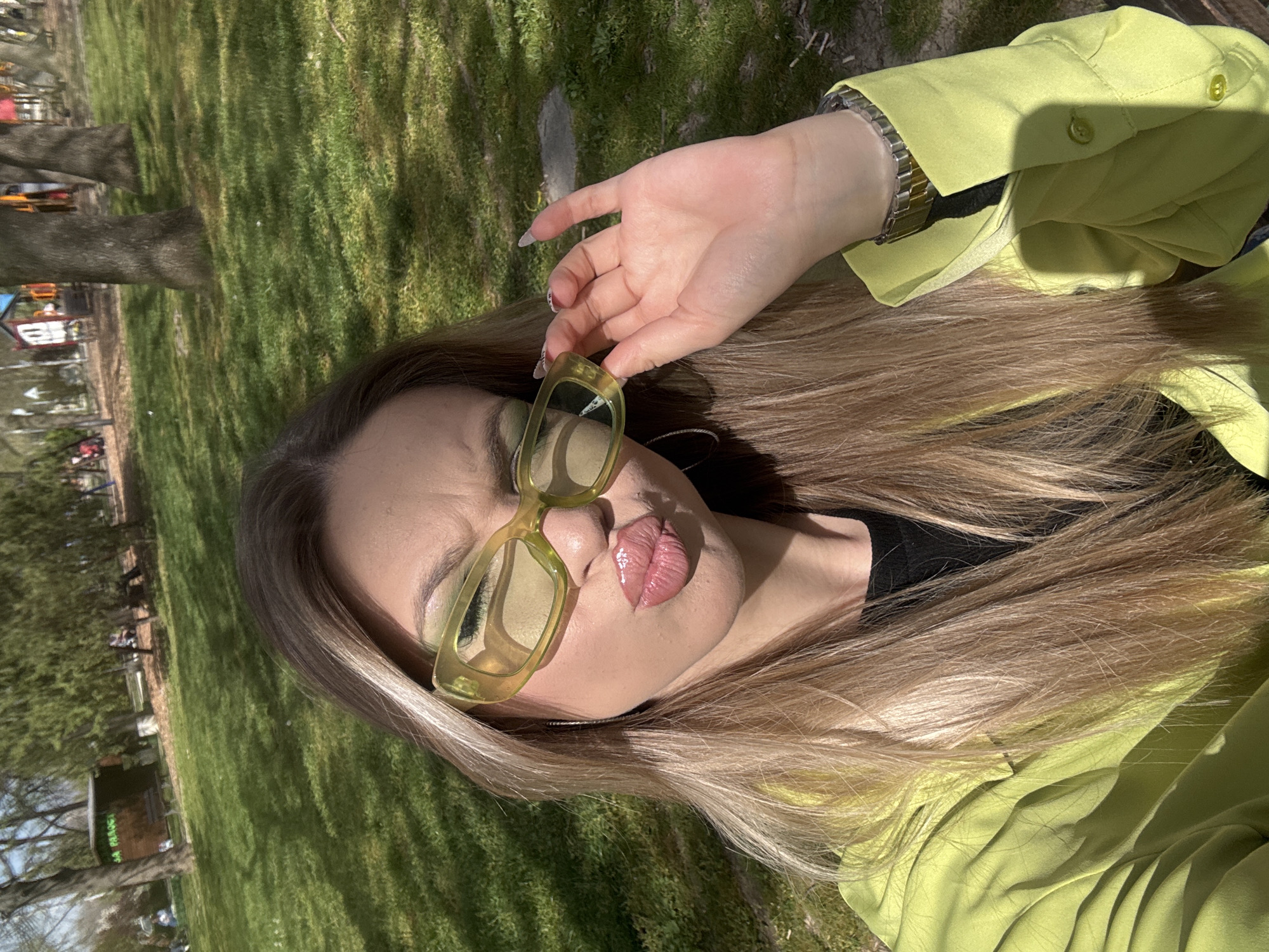A woman is captured in a close-up, angled outdoor shot, posing under bright sunlight in what appears to be a park in Plovdiv, Bulgaria. She has long, light brown hair that cascades over her shoulders and upper body. She wears a vibrant lime green long-sleeved shirt or jacket, with a metallic watch visible on her left wrist. Her right hand is raised, gently holding a pair of distinctive light yellow, thick-framed sunglasses, which she holds near her face, partially obscuring her eyes. Her lips are glossy and slightly parted.
The background consists of a well-lit grassy area with trees casting shadows, indicating a sunny day. In the far background to the left, colorful structures resembling playground equipment are visible, along with a small brown building. Indistinct green text can be vaguely seen on this building. The overall mood is relaxed and stylish, suggesting a leisurely moment in a public green space.