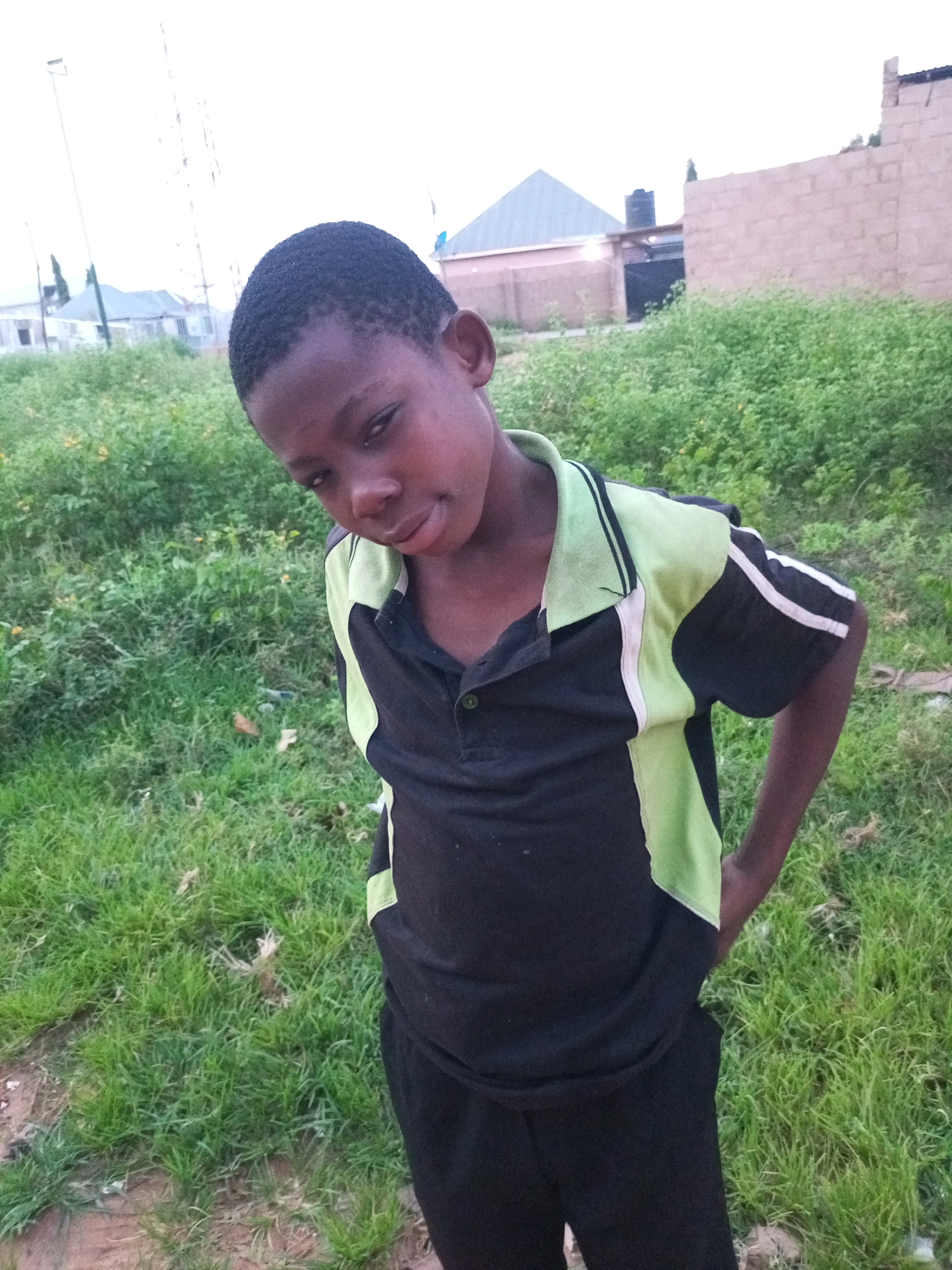 The image shows a young boy standing in an outdoor, grassy area. He is wearing a black polo shirt with lime green accents and black pants. His hands are in his pockets, and he is looking towards the camera with a slight smirk. The boy has dark skin and short, dark hair.
The background is filled with lush green grass and foliage, with some buildings and structures visible in the distance. The sky is overcast, suggesting it might be late afternoon or early evening, or perhaps cloudy weather. The scene appears to be in a rural or semi-urban setting in Tempure, Nigeria, as indicated by the buildings and the general landscape. There is no visible text in the image. The boy's expression is relaxed and somewhat playful.