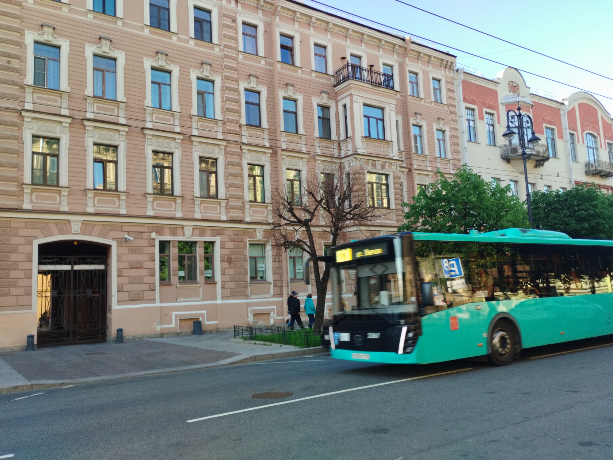 The image depicts a street scene in Saint Petersburg, Russia, with a prominent, pale pink building lining the left side of the frame. The building exhibits a classical architectural style with decorative window surrounds and a mansard roof. On the ground floor, a large arched entryway with an ornate metal gate is visible. A modern, teal-colored bus is captured in motion, partially obscuring the building and street. The bus is branded with Cyrillic text and a route number displayed on its front. Two individuals are walking on the sidewalk in front of the building, and a tree with bare branches stands between them and the bus. The street is paved, and overhead wires suggest the presence of public transportation. The sky is clear and blue, indicating daytime.