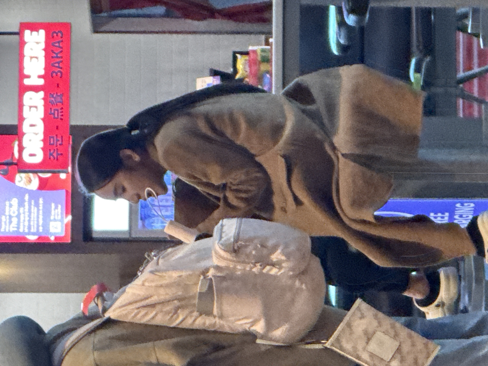 The image depicts a person, likely female, with dark hair pulled back in a ponytail, sitting on a stool and holding a white object to their mouth. They are wearing a tan coat. In the background, a red sign with white lettering reads "ORDER HERE" in English, and also in Korean and Chinese. To the left of the person, a light-colored backpack with multiple pockets is visible. The environment appears to be an indoor commercial setting, possibly a food establishment, with a counter or table in front of the person. The lighting is somewhat dim, with a bright advertisement screen visible behind the ordering sign. The setting does not provide specific visual cues to identify the exact location within Bắc Cam Ranh, Vietnam.