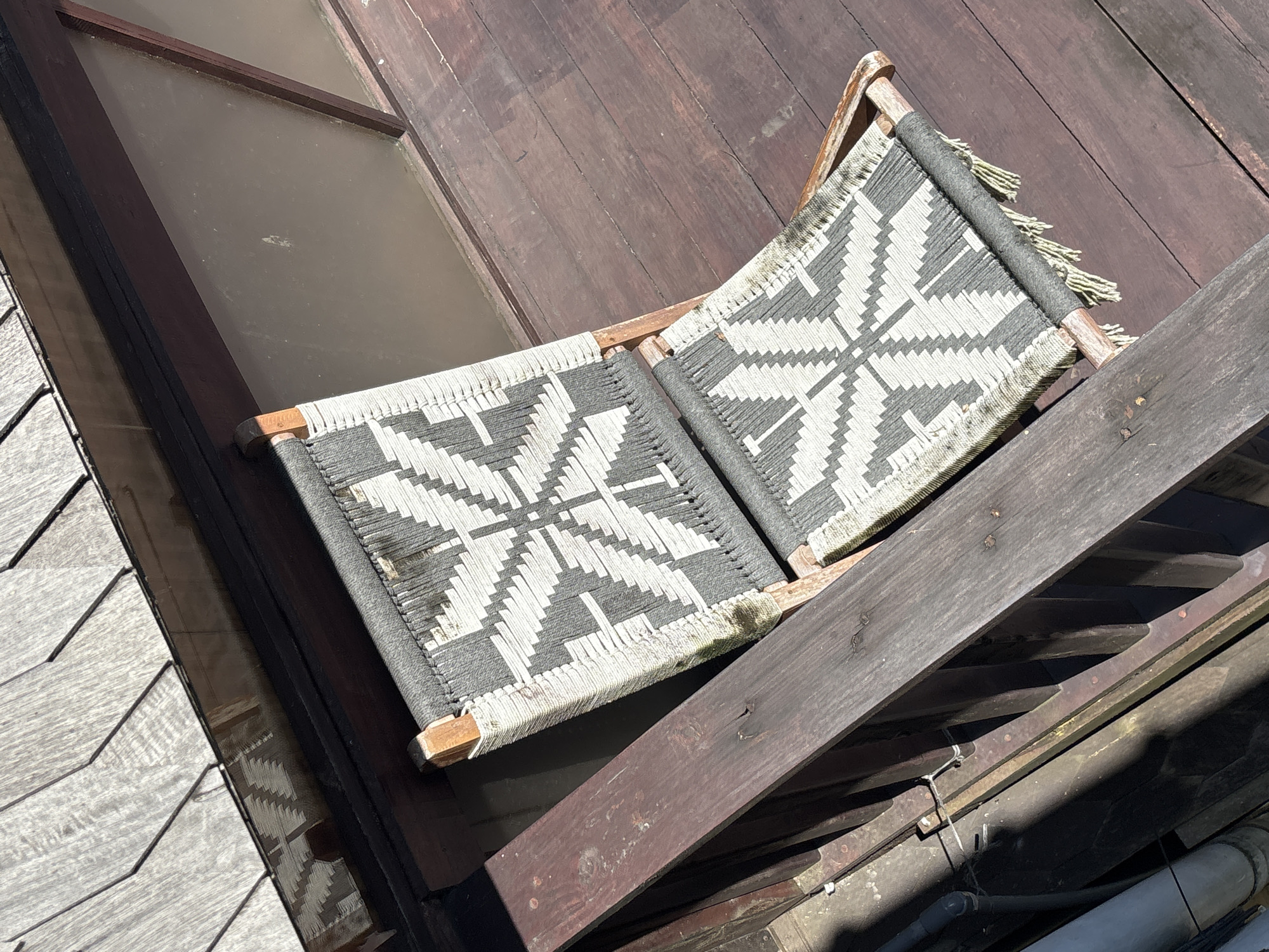 An outdoor scene captured from an elevated perspective shows a traditional, possibly handmade, wooden folding chair with a woven seat and backrest. The weaving features a geometric pattern in white and grey threads, creating star-like or snowflake designs. The chair's frame is made of light-colored wood, and some of the weaving ends are visible as tassels.
The chair is resting on or next to various wooden surfaces. To its left, a structure composed of overlapping light-grey wooden planks, possibly a roof or wall, is visible. To its right and beneath it, darker brown wooden planks, resembling decking or flooring, dominate the background.
A large rectangular translucent panel or sheet of glass, set within a dark wooden frame, occupies a significant portion of the upper left quadrant. This panel appears to be part of a roof or floor, allowing light to pass through. Below the chair, towards the bottom right, some dark wooden beams, metal brackets, and a few pipes or tubes are partially visible, suggesting underlying structural or utility elements.
The scene is brightly lit by direct sunlight, casting strong shadows, which indicates it is midday or early afternoon on a clear, sunny day. No people or activities are present. The setting appears to be a rustic or traditional outdoor space, possibly a deck, balcony, or rooftop, consistent with its location in Pekutatan, Indonesia. There is no visible text in the image.