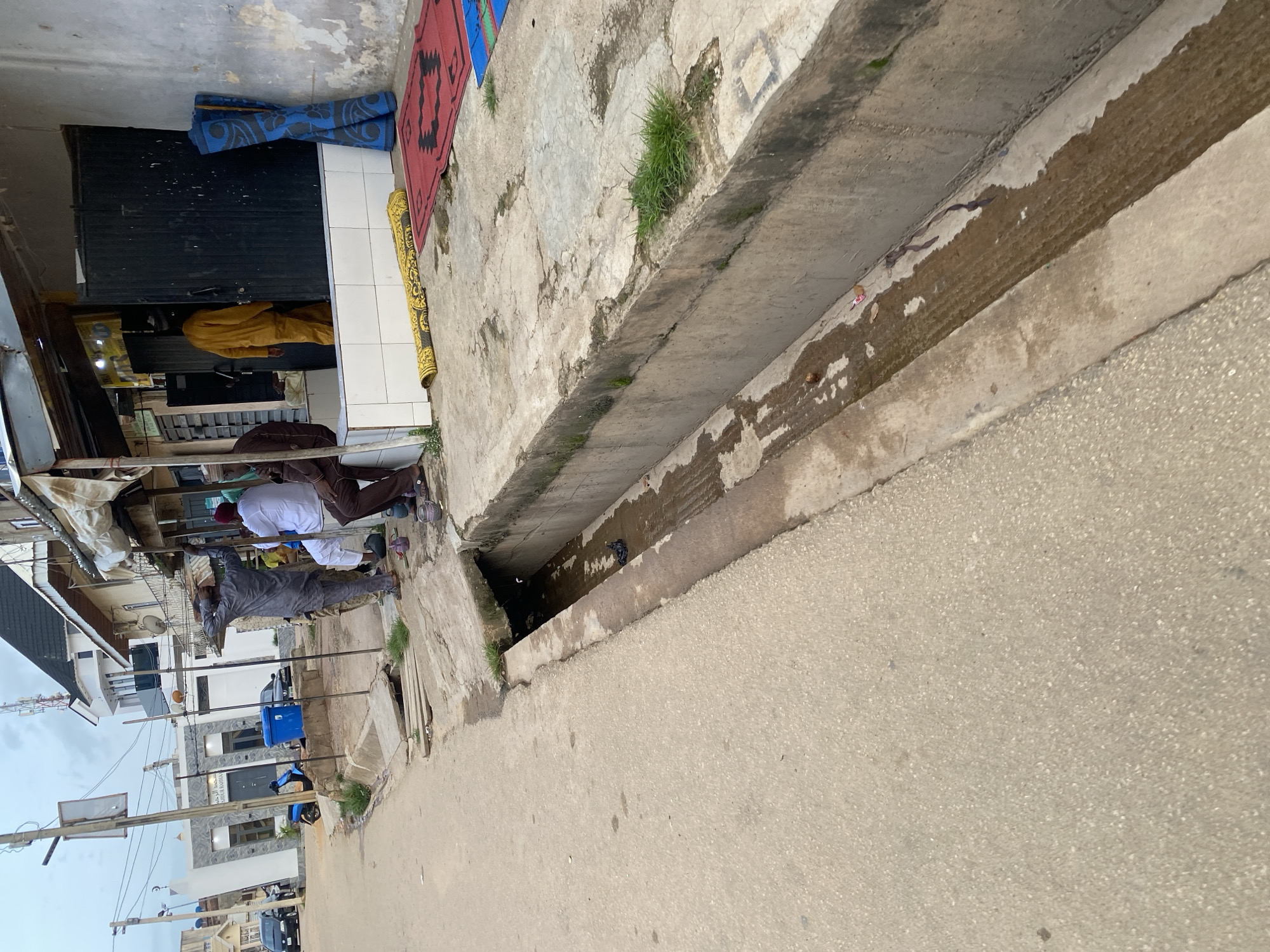A street scene in Jos, Nigeria shows a concrete drainage ditch running alongside a paved road. Several people are gathered near a storefront. One man in a grey traditional outfit stands with his hands on his head, facing away from the camera. Two other men in white and brown traditional attire are seated near him. A fourth person in a yellow outfit is walking into the storefront. The storefront has a black door and an awning. Mats, one red and two blue, are stacked vertically outside the storefront. Another yellow mat is laid out on the ground. The sky is overcast.