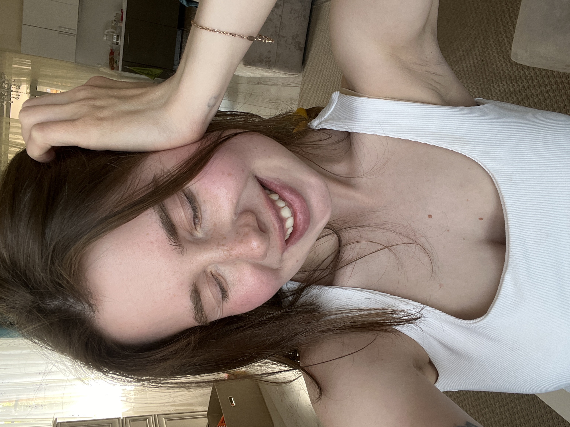 A young adult female with long, brown hair, a fair complexion, and visible freckles across her nose and cheeks is depicted in a close-up, high-angle self-portrait. Her eyes are closed, and she displays a wide, open-mouthed smile, revealing her upper teeth and creating rosy cheeks. She is wearing a white, ribbed tank top with wide straps and a square neckline; a glimpse of a lighter, possibly cream-colored undergarment is visible beneath the neckline. Her right hand is raised, with fingers splayed, resting on the top of her head amidst her hair. A rose gold-colored chain bracelet, adorned with small charms (including a star, a cross, and a circular element), is visible on her right wrist. The area of her left armpit is partially visible, showing natural skin texture.
The background suggests an indoor, residential setting, likely an apartment or house, illuminated by bright natural light from a window to the left, which has sheer curtains. To the left, a light-colored, multi-drawer metallic piece of furniture is partially visible, along with a brown cardboard box on the floor. The right side of the background reveals light-colored flooring, a beige rug, and the corner of a light-colored upholstered sofa or armchair. Further back, through an opening, a kitchen area is visible with white upper cabinets, darker lower cabinets, a countertop, a sink, and several kitchen items including a kettle. A chandelier hangs from the ceiling in the background.