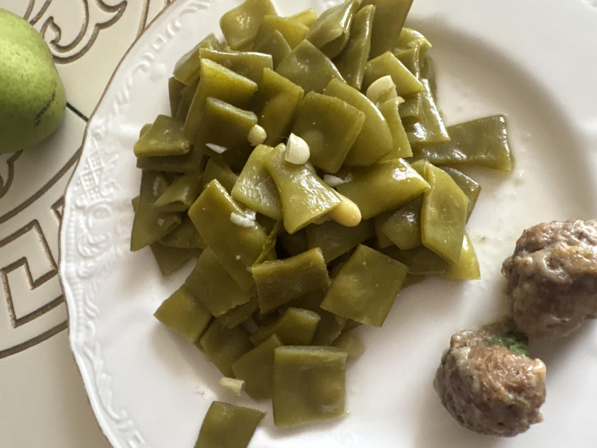 In Georgiyevsk, Russia, a white plate with a decorative rim sits on a patterned surface. The plate is filled with chopped green beans, interspersed with pieces of garlic. To the bottom left of the plate are two meatballs coated in a light sauce. In the upper right corner of the image, a green pear rests on the patterned surface, partially visible. The lighting suggests an indoor setting.