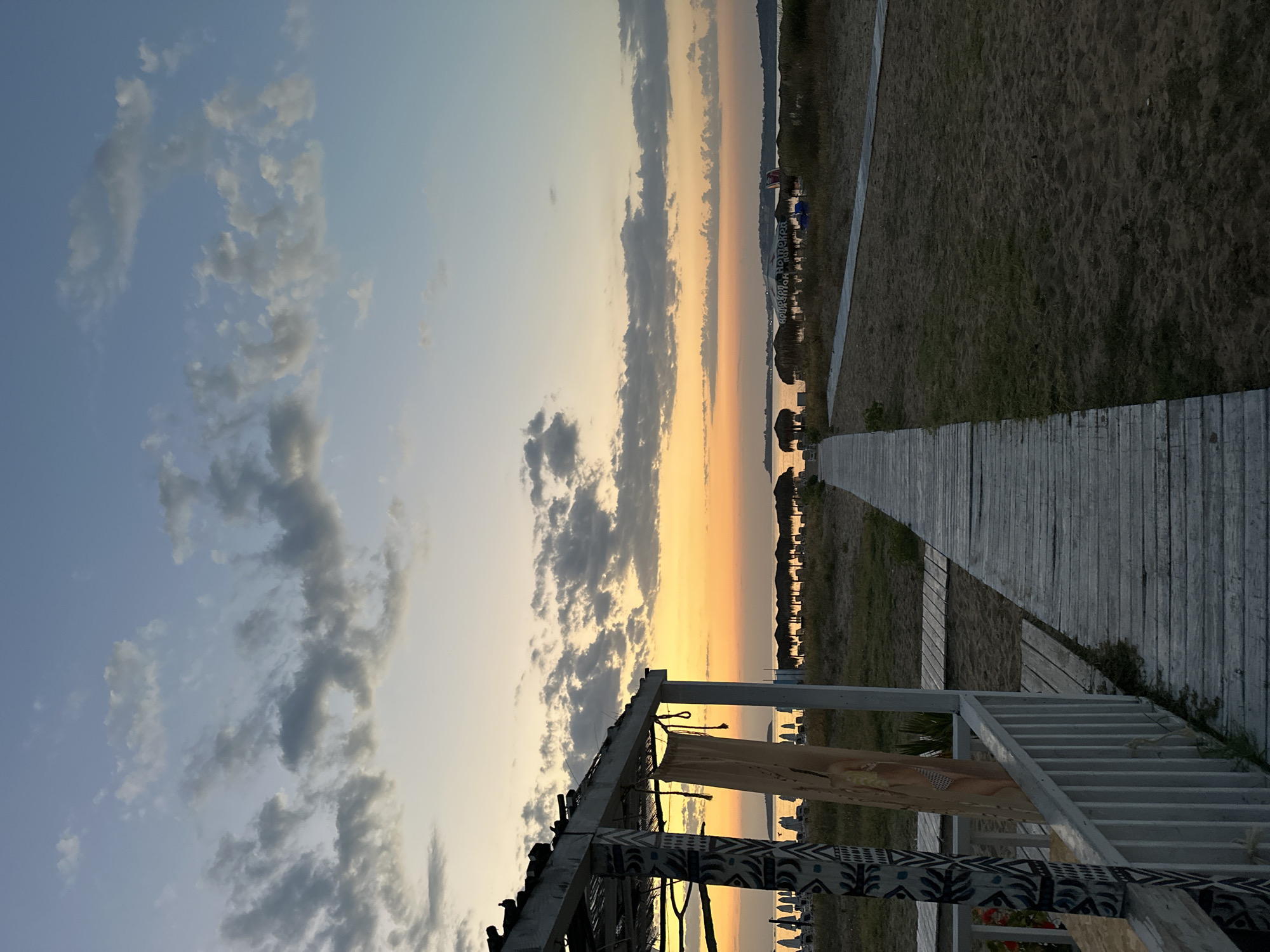 A weathered wooden boardwalk leads across a sandy beach toward a calm sea at sunset. The sky is painted with hues of orange and yellow near the horizon, transitioning to a soft blue with scattered grey clouds. On the left, a white wooden structure with decorative patterns on its pillars provides shade. In the distance, rows of thatched umbrellas and lounge chairs are visible along the coastline. The scene is tranquil and devoid of people. The location is on the beach in Sozopol, Bulgaria, facing the Black Sea.