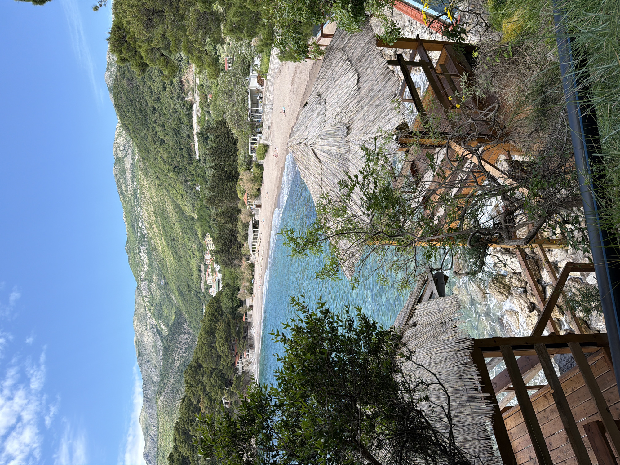 The image depicts a coastal scene in Petrovac centar, Montenegro. A beach with tan sand slopes gently to turquoise water. A mountain with lush vegetation rises on the left. A man-made structure is prominent in the foreground, possibly a partially obscured wooden platform or shelter with a thatched roof. The platform is surrounded by greenery. The sky is light blue with scattered clouds. The image is taken from an elevated perspective, looking down towards the beach and the Adriatic Sea. People are visible on the beach, suggesting it's a tourist area. The vegetation is thick and comprises a mixture of trees and shrubs, typical of a Mediterranean coastal environment.