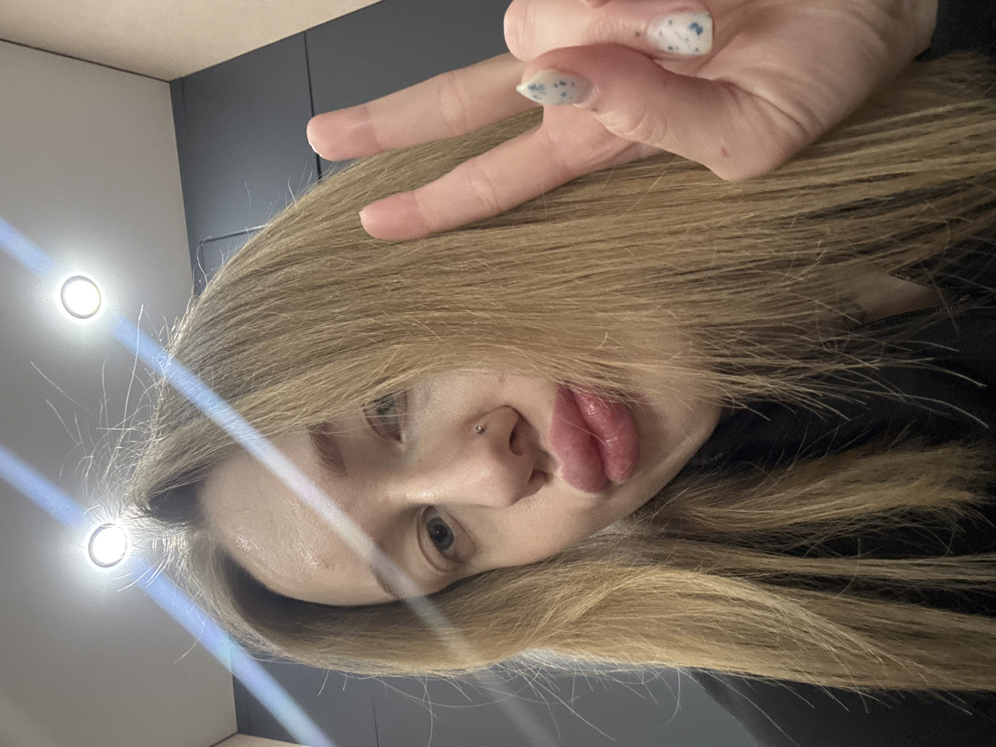 A rotated close-up selfie shows a young woman with long, blonde hair partially covering her face, making a peace sign with her right hand. She has a piercing on the left side of her nose. The background appears to be an indoor setting with light fixtures visible in the upper left corner. The image does not depict any specific location in Azov, Russia or contain any information to suggest that location.